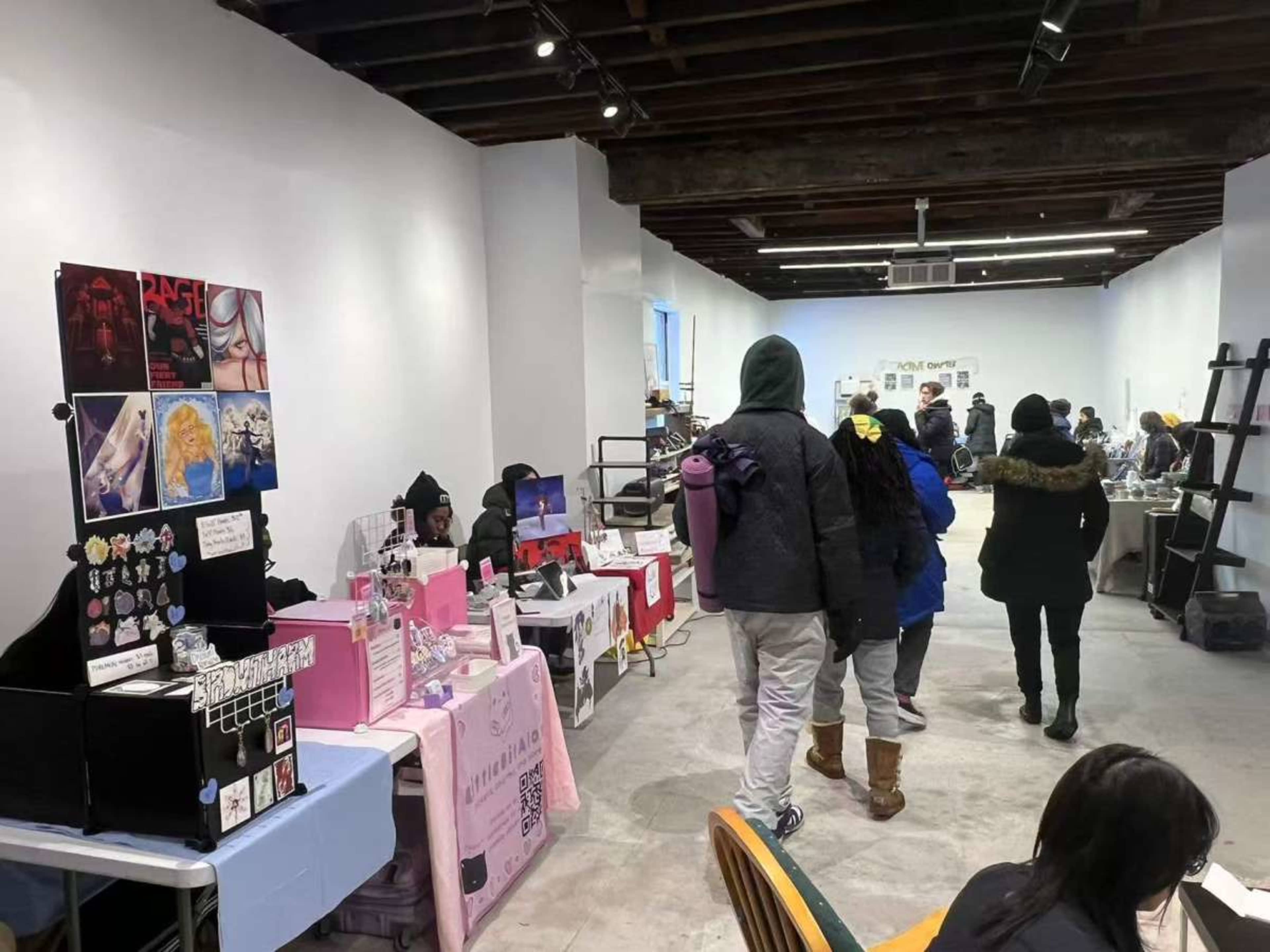 The image shows a bustling market or exhibit space with various tables displaying art and merchandise, and visitors browsing the products while wearing winter clothing.
