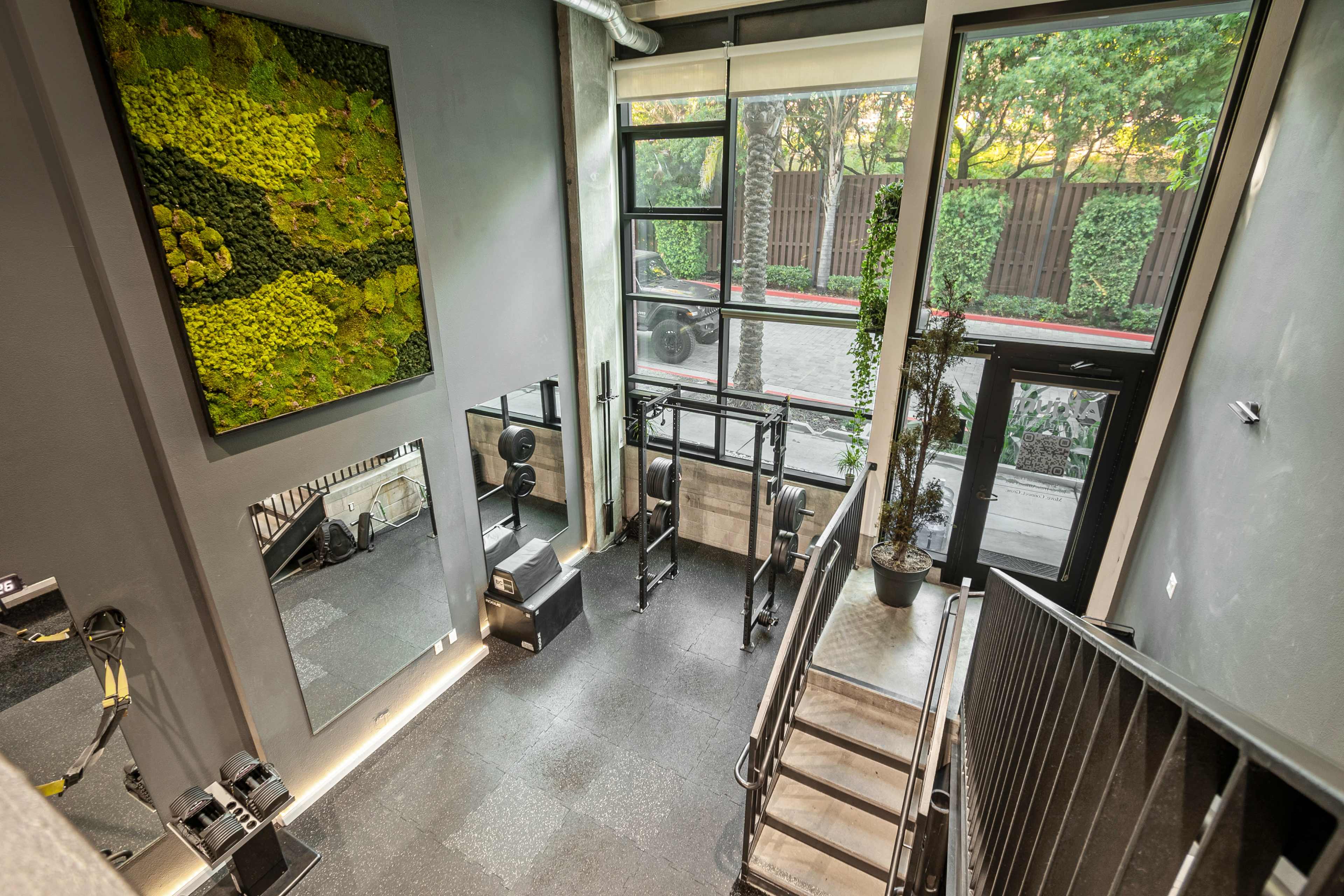 A modern fitness space with large windows, a staircase, workout equipment, and a moss wall art installation.