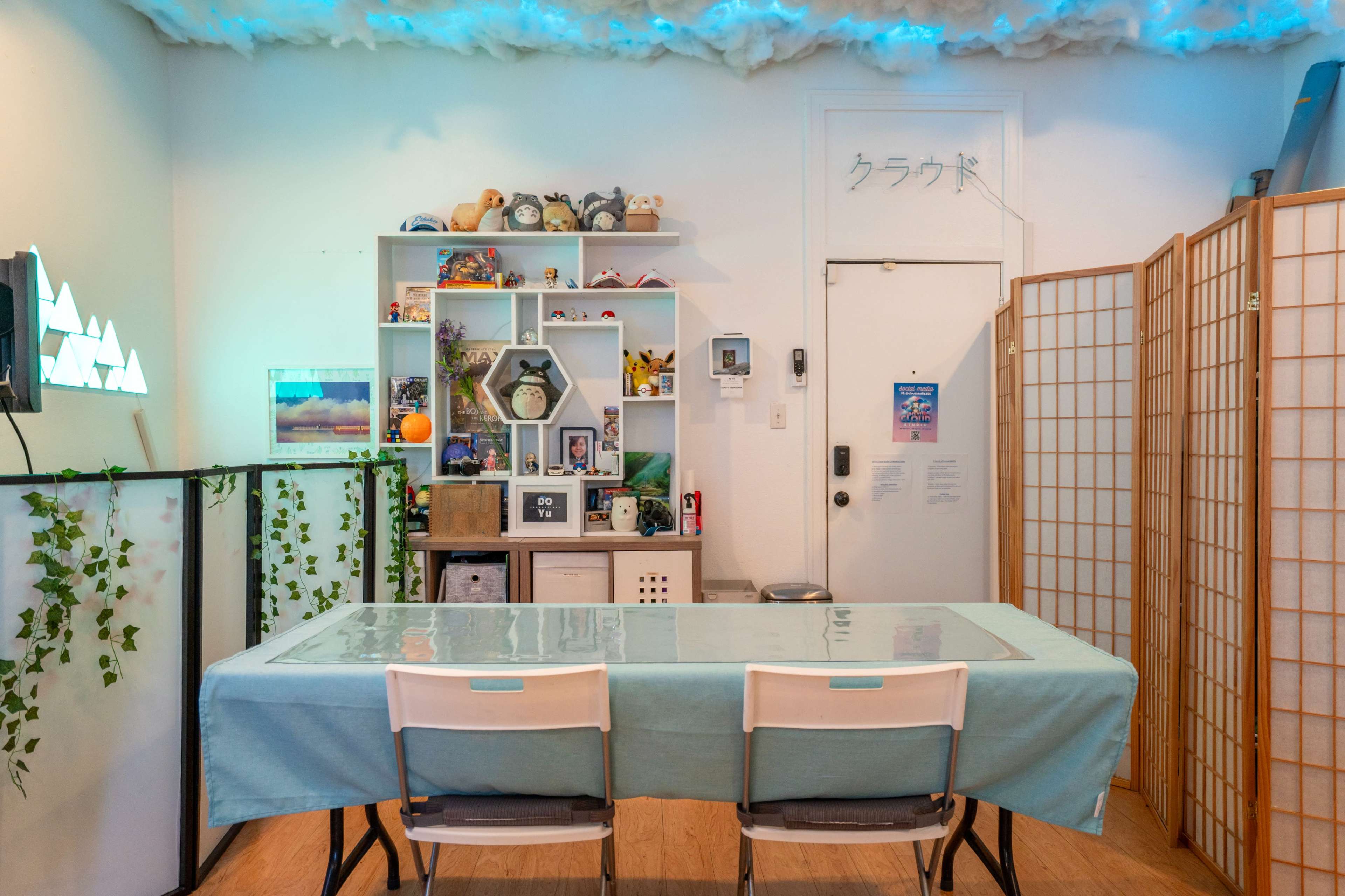 The image shows a brightly lit room with a large table, surrounded by chairs, and decorated shelves filled with various items against a backdrop of soft cloud-like fabric on the ceiling.