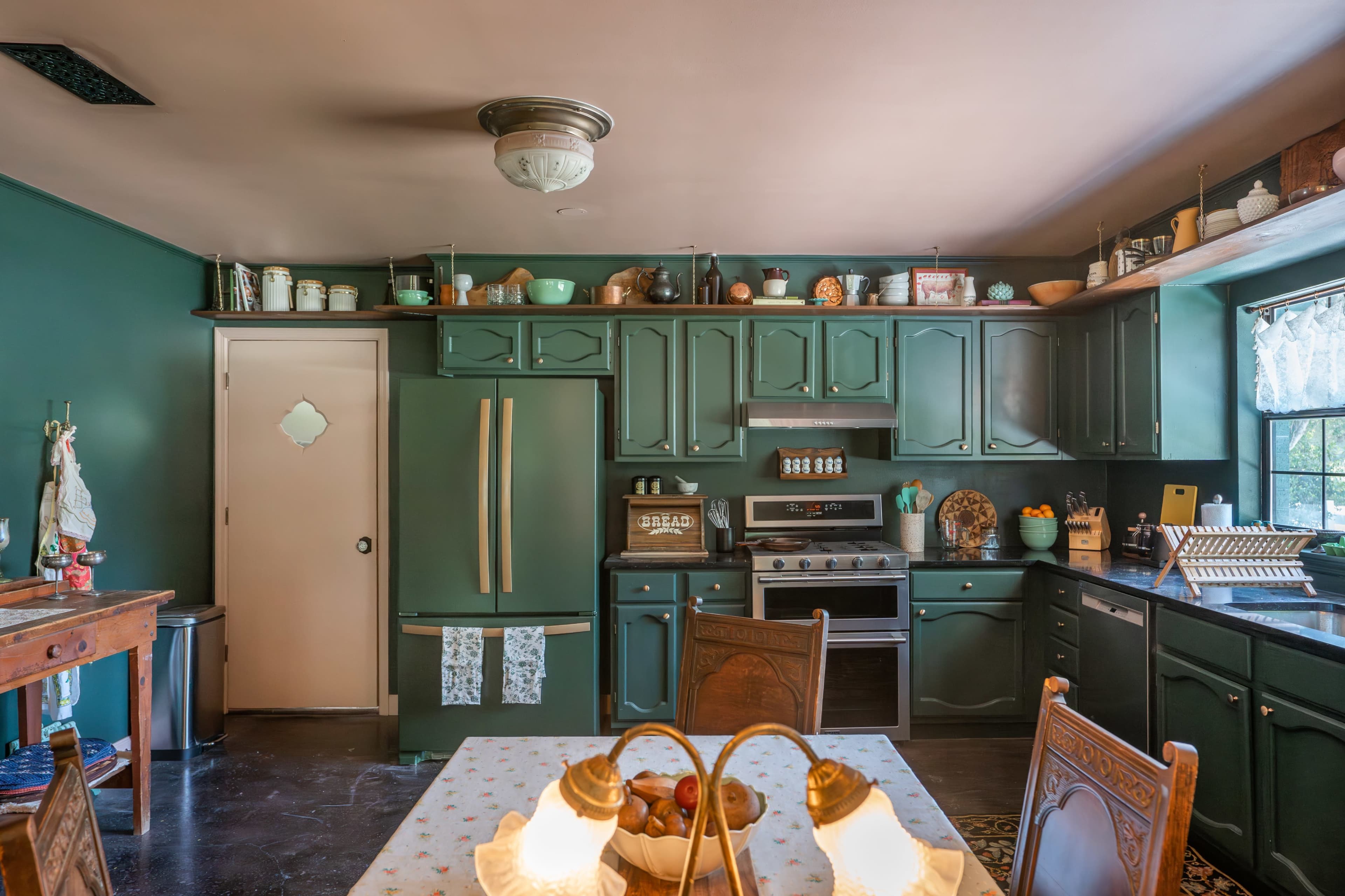 The kitchen features green cabinetry, a stainless steel refrigerator, a stove, a dining table with two lamps, and shelves filled with various kitchenware.