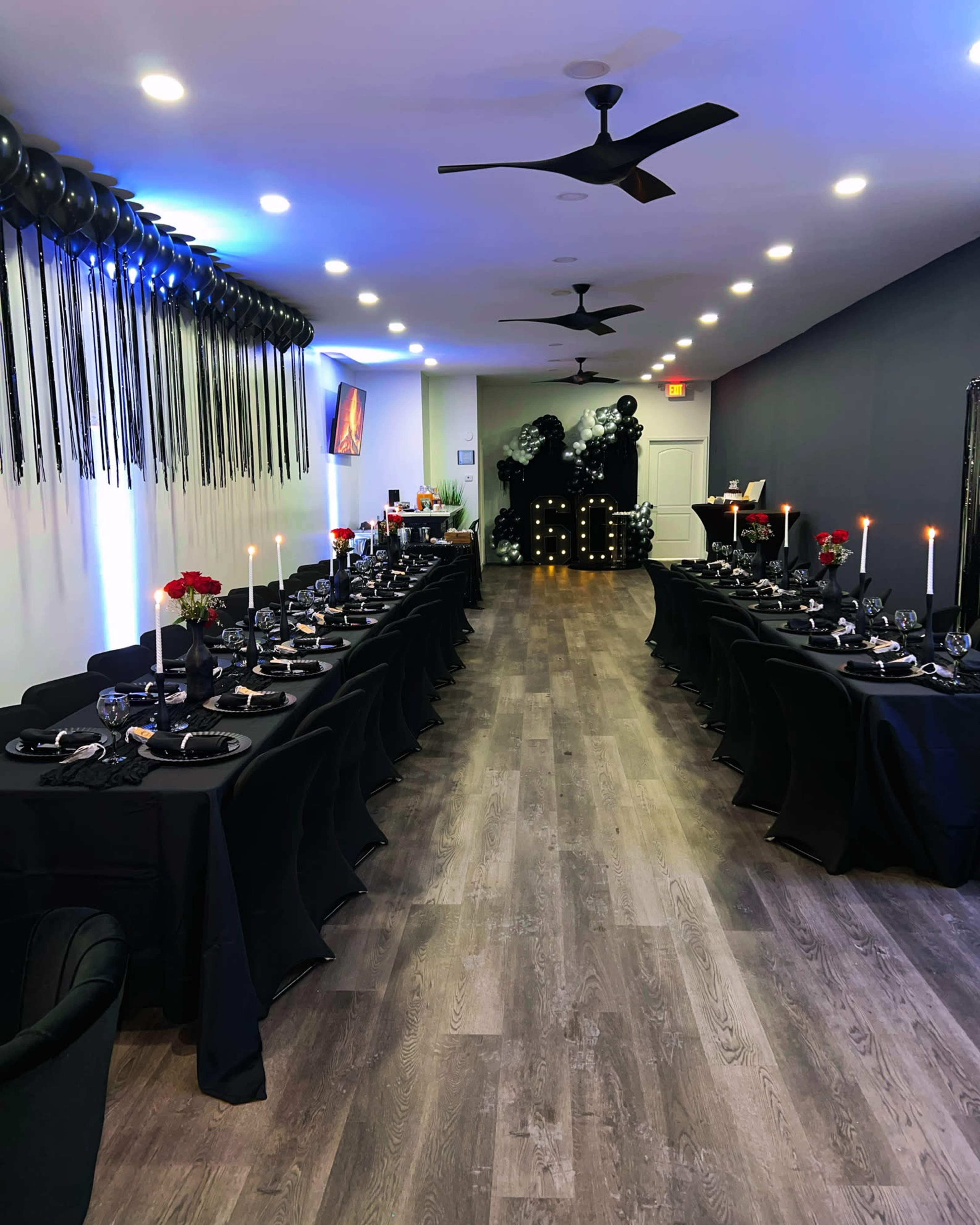 A long, elegantly set dining table is arranged in a dimly lit room with black tablecloths, candles, and decorative balloons along the walls.