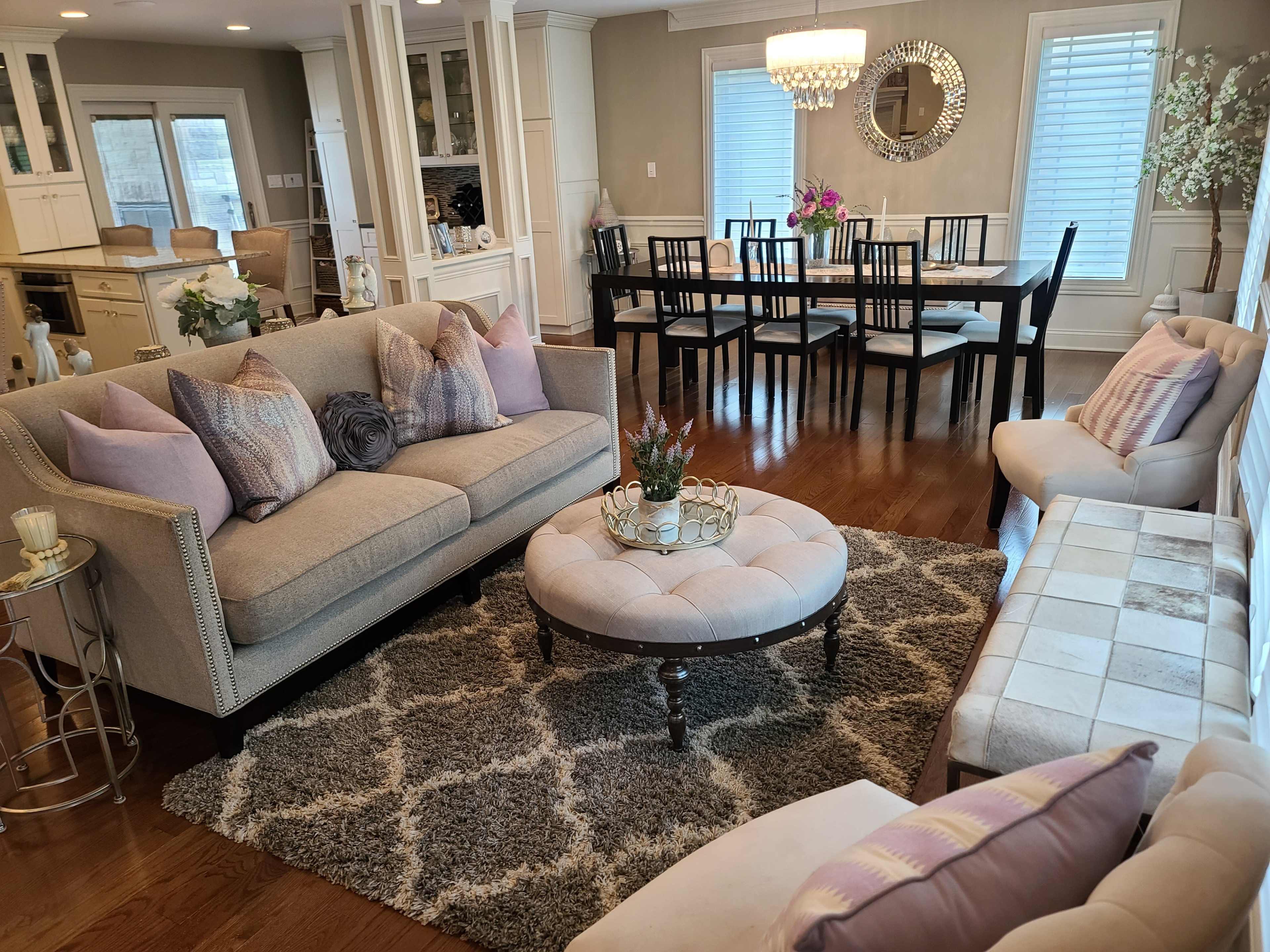 The living area features a gray sofa and a round ottoman on a plush rug, with a dining table set for six in the background.