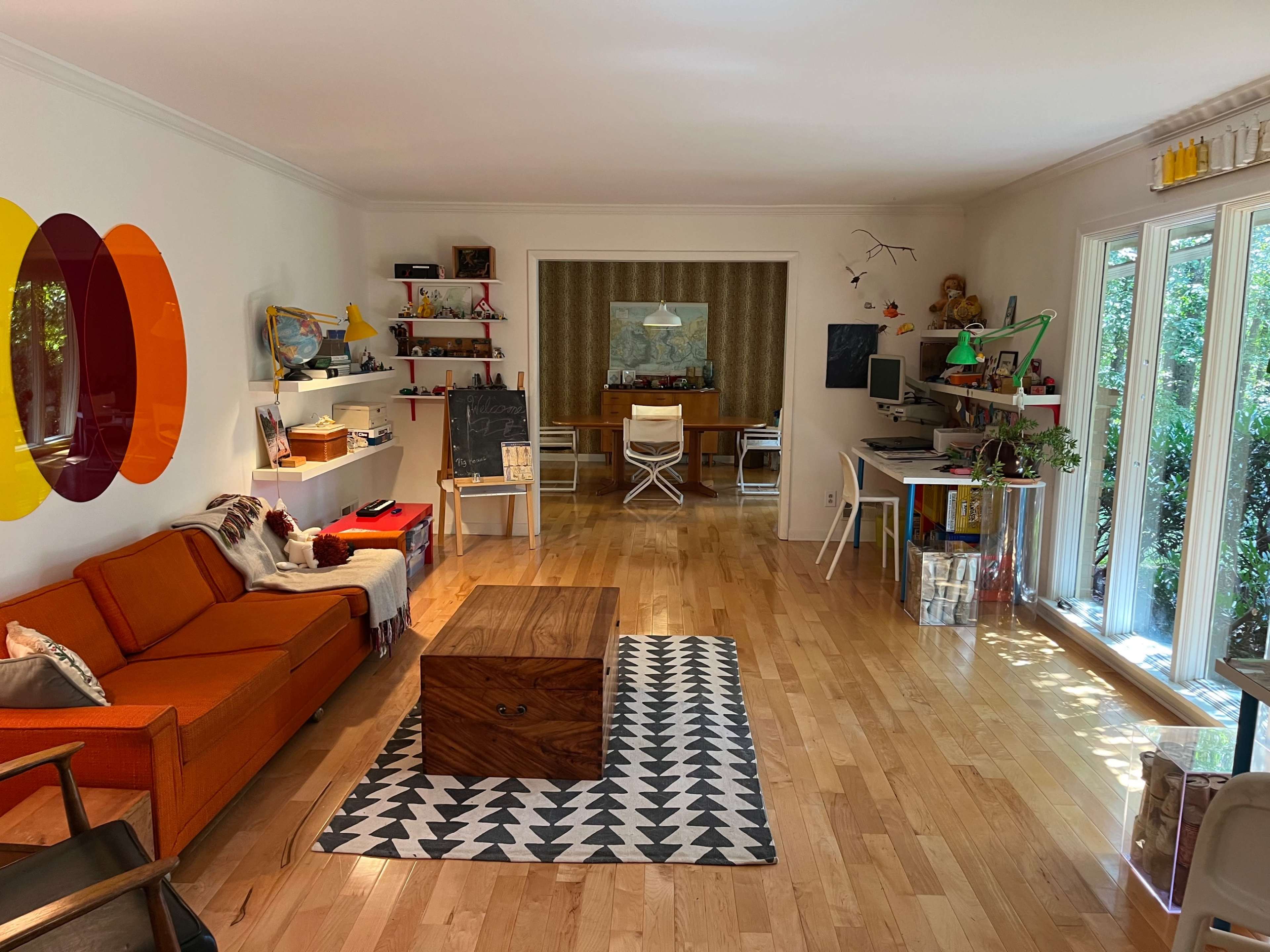 The image shows a spacious living area with a bright orange sofa, a wooden coffee table, and a modern workspace in the background surrounded by large windows.