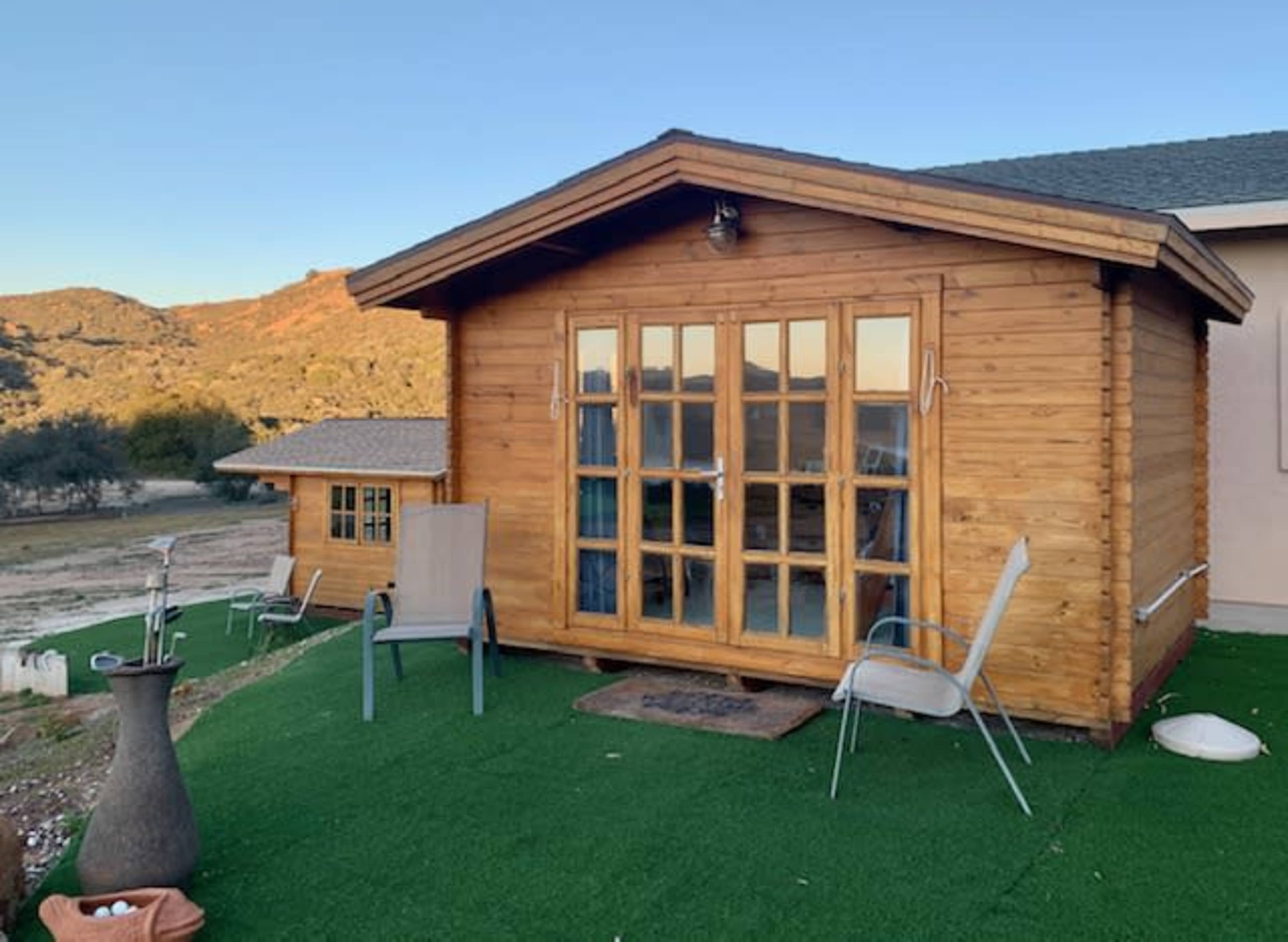 A wooden cabin with large front windows is situated on a grassy area next to a hillside in the background.