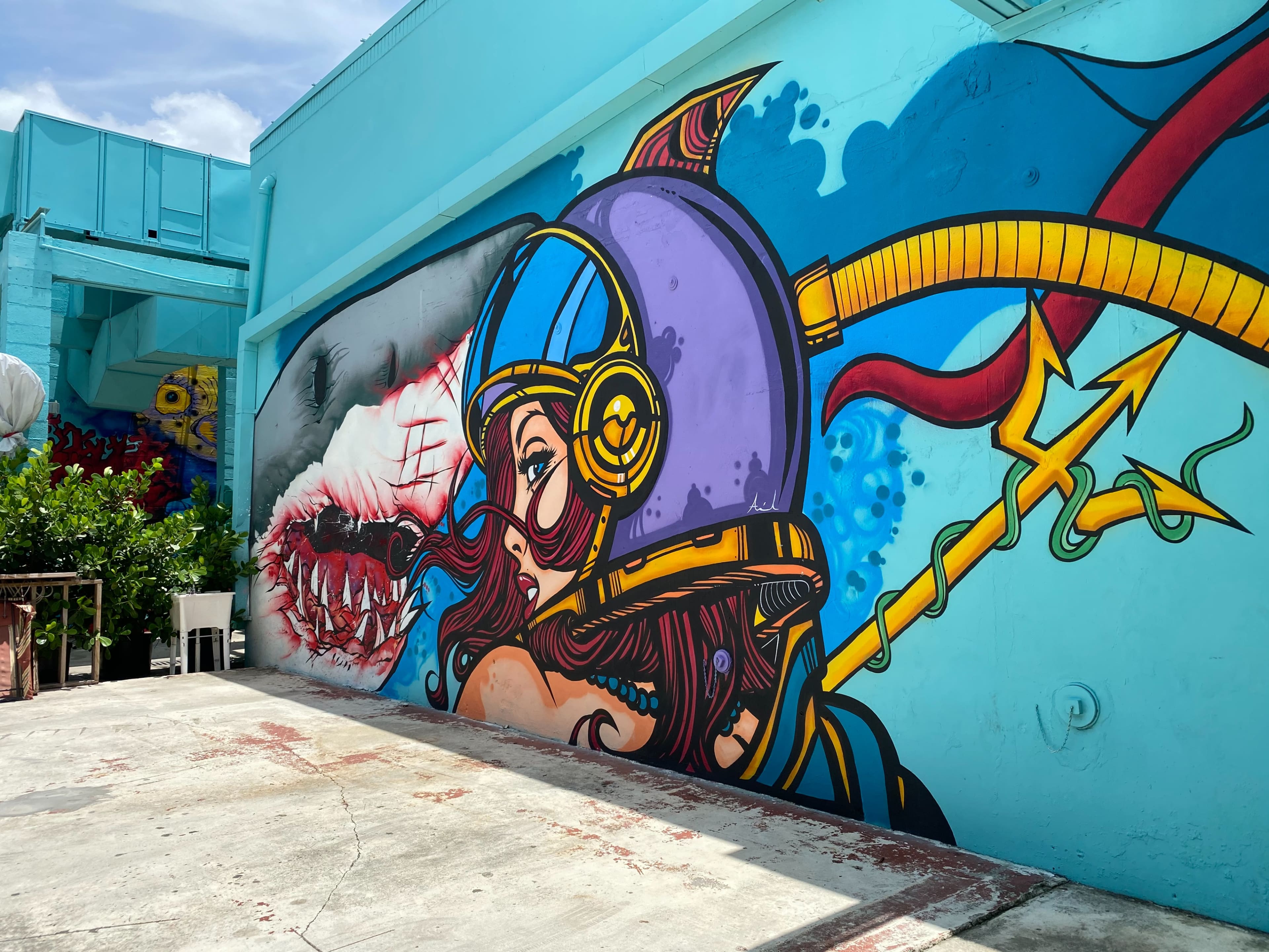 A vibrant mural featuring a woman in a diving helmet alongside a large, artistic shark is painted on a bright blue wall.