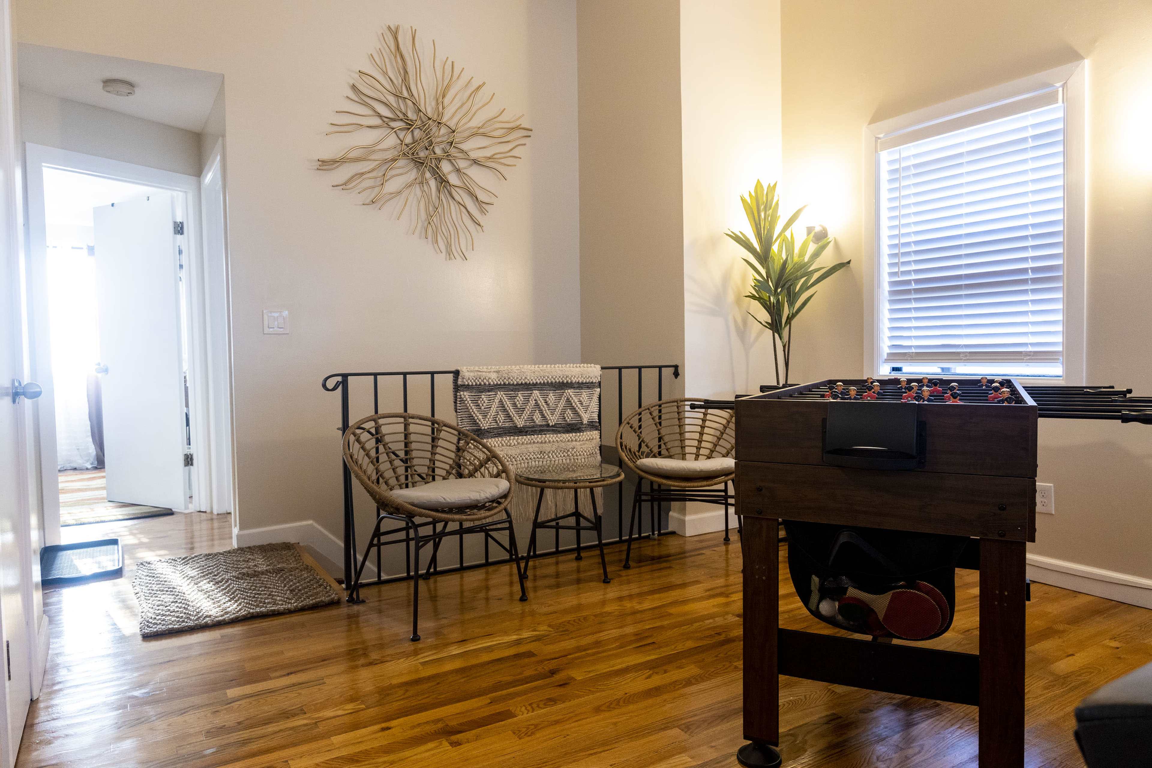 A room with a foosball table in the foreground, two wicker chairs and a decorative wall piece, with a doorway leading to another room in the background.