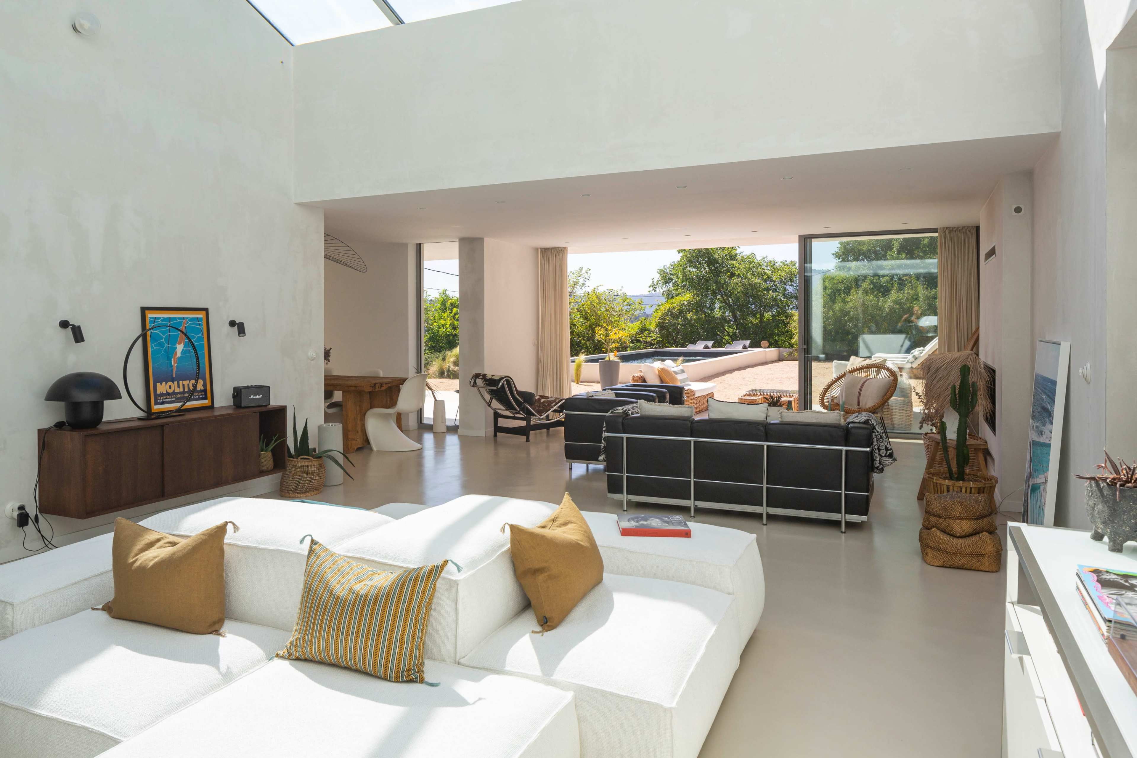 The image shows a modern, open-concept living space featuring a white sectional sofa, a wooden dining table, and large glass doors that lead to an outdoor area.