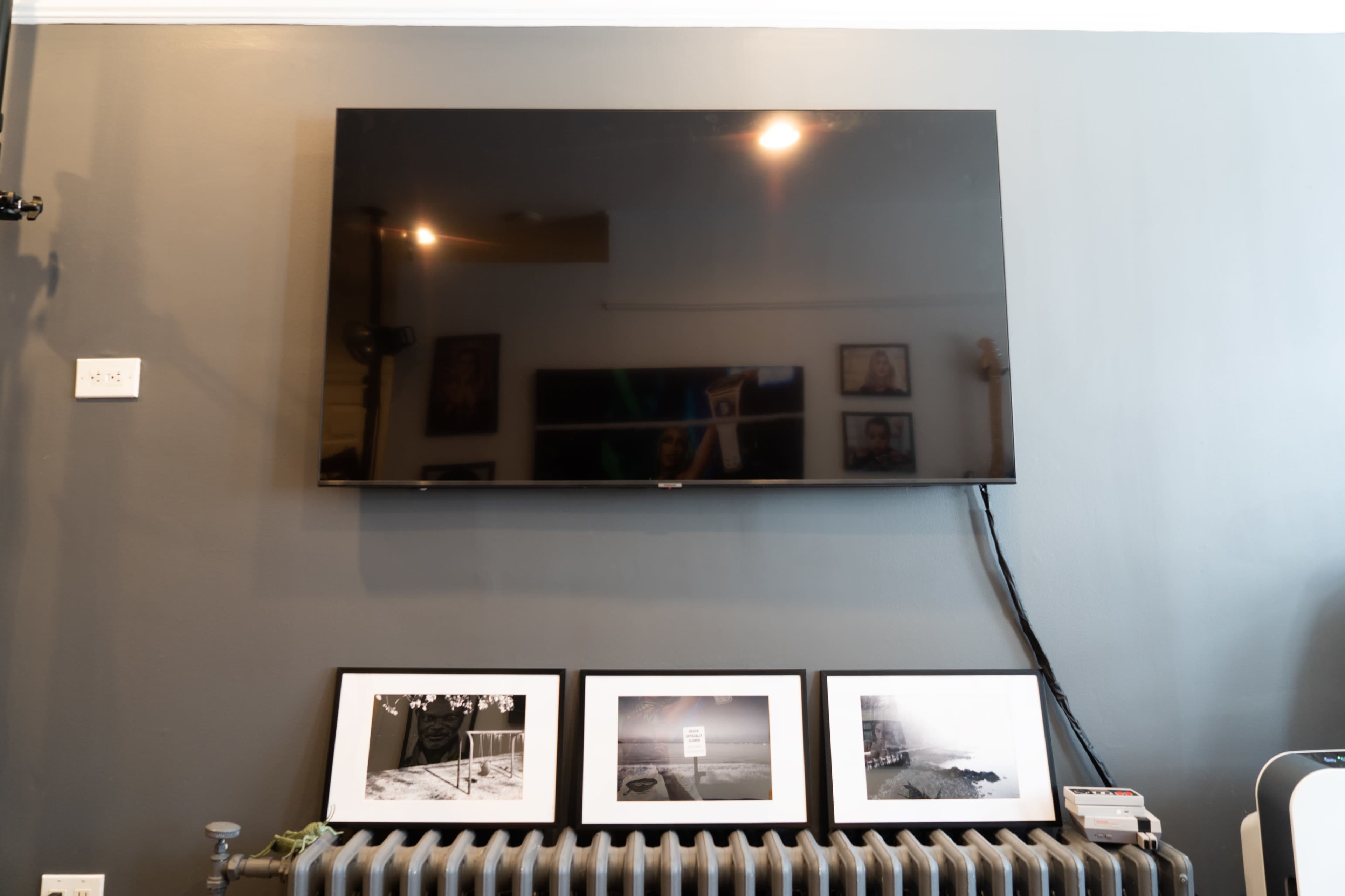 A flat-screen television is mounted on a gray wall above four framed photographs displayed on a radiator.