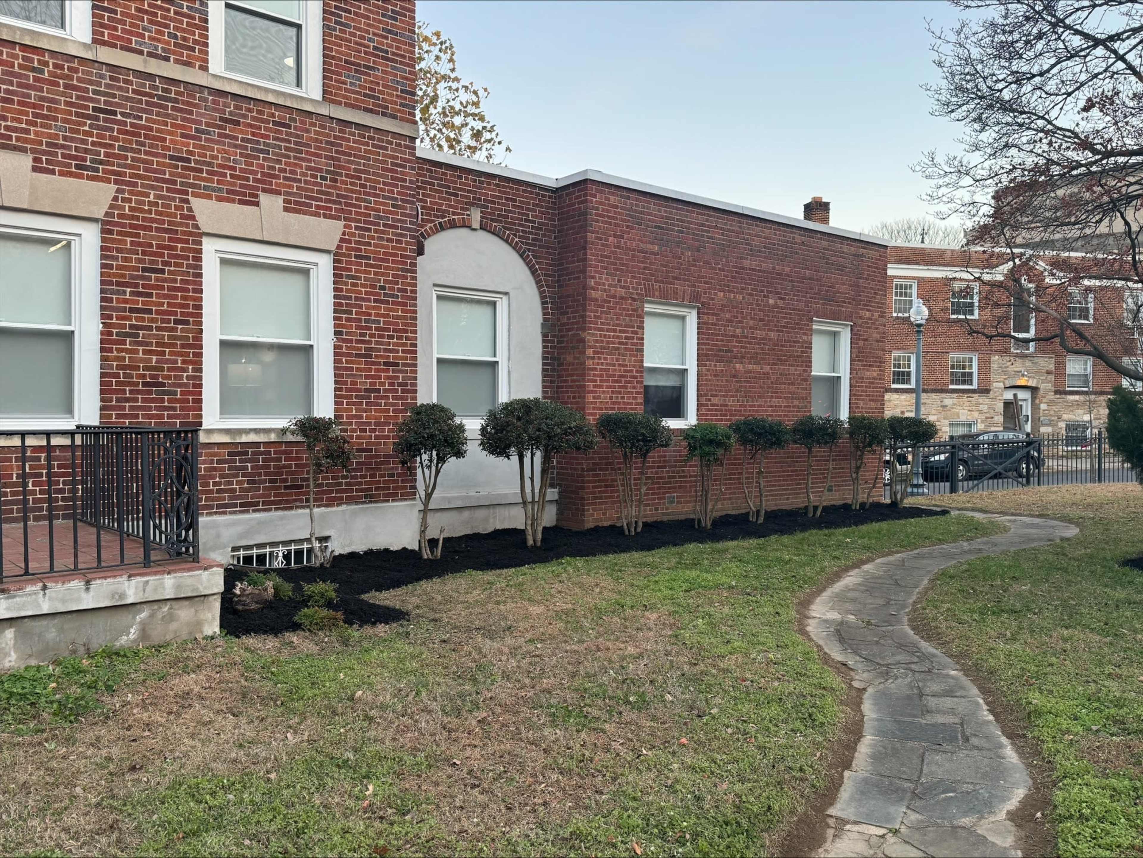 The image shows a landscaped area beside a brick building, featuring a curved pathway and neatly trimmed shrubs along the foundation.
