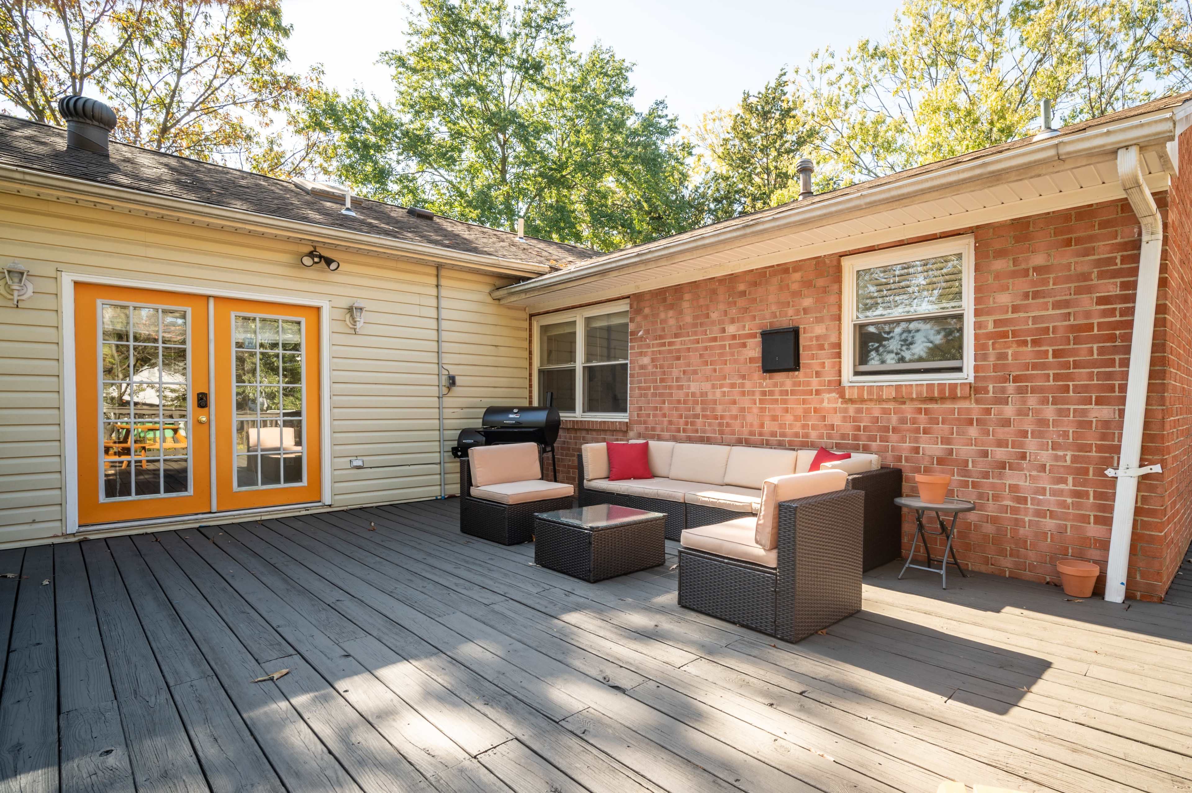 The image shows a wooden deck with outdoor seating, a grill, and sliding glass doors leading into a house.