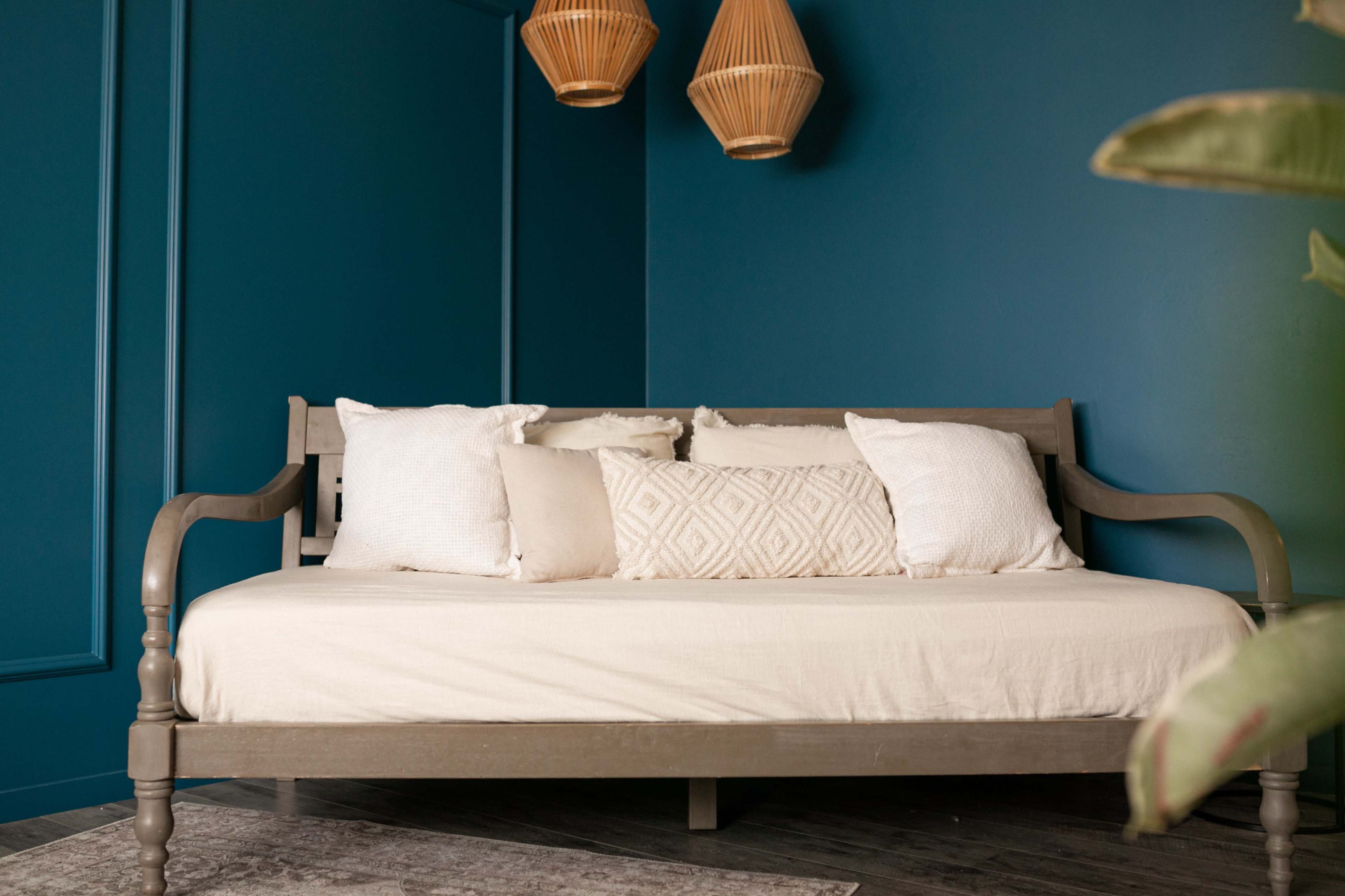 A wooden daybed with beige cushions is positioned against a dark blue wall, illuminated by woven pendant lights.