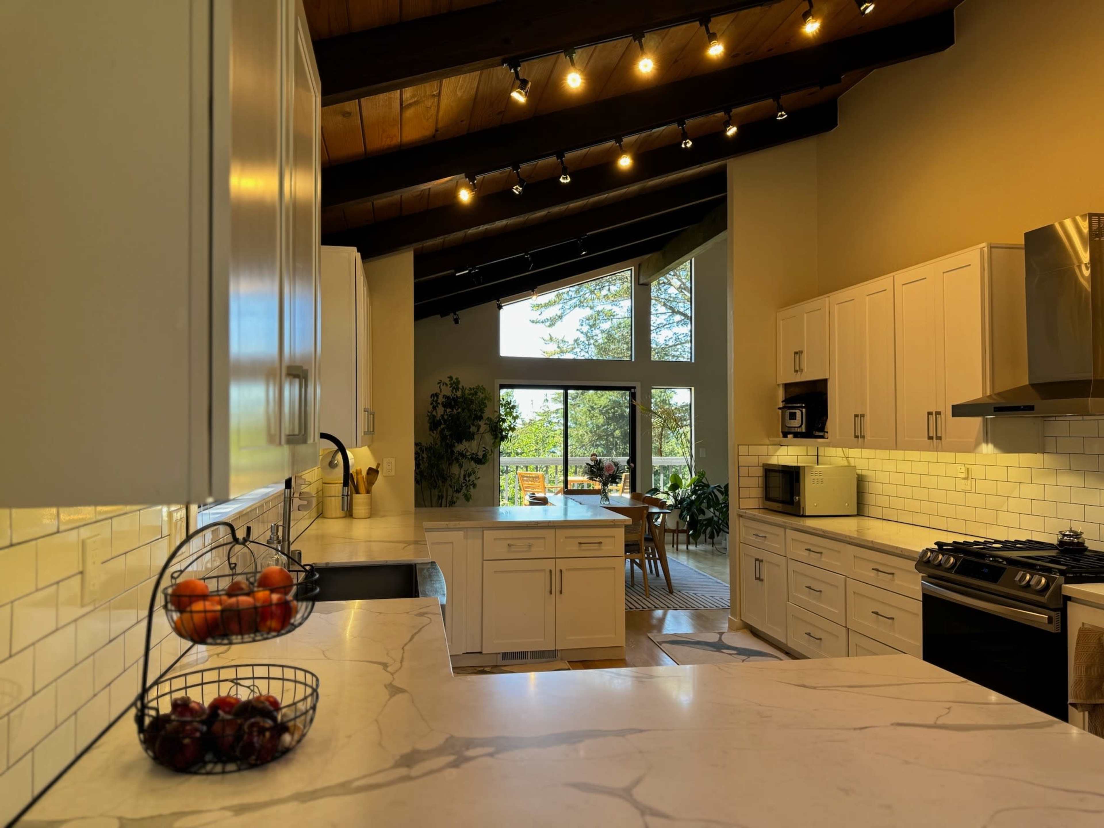 A modern kitchen with white cabinetry, a marble countertop, and large windows overlooking a green outdoor area.