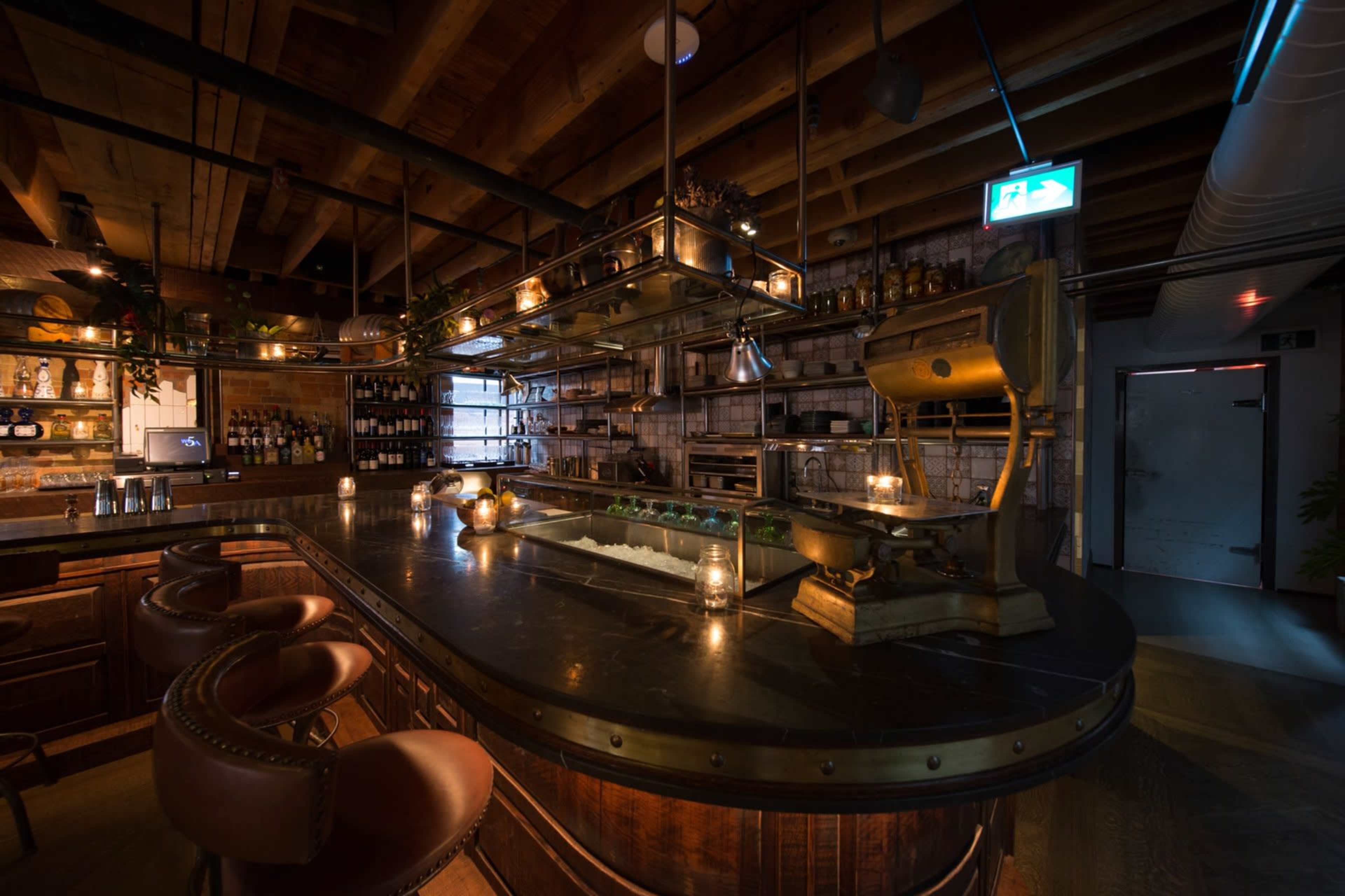The image shows a dimly lit bar with wooden beams, a curved counter, and an array of shelves stocked with bottles and kitchenware.
