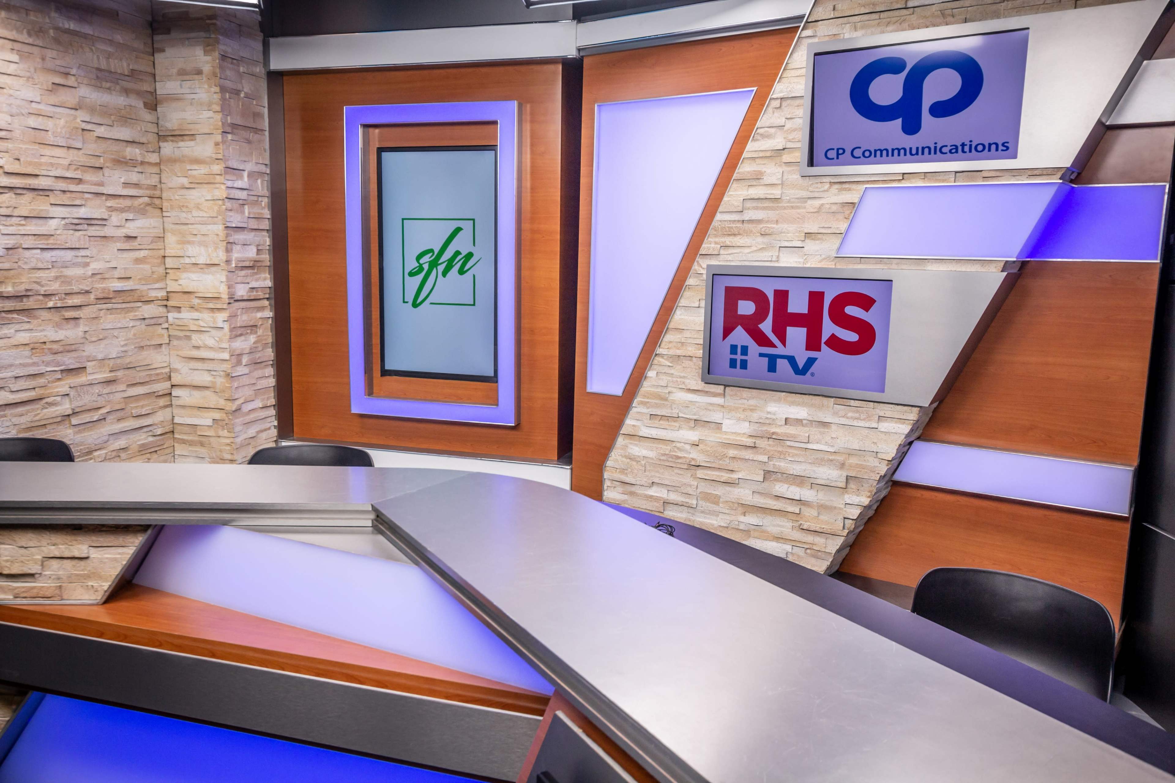 A modern television studio set featuring logos for "CP Communications" and "RHS TV" against a background of wood paneling and illuminated accents.