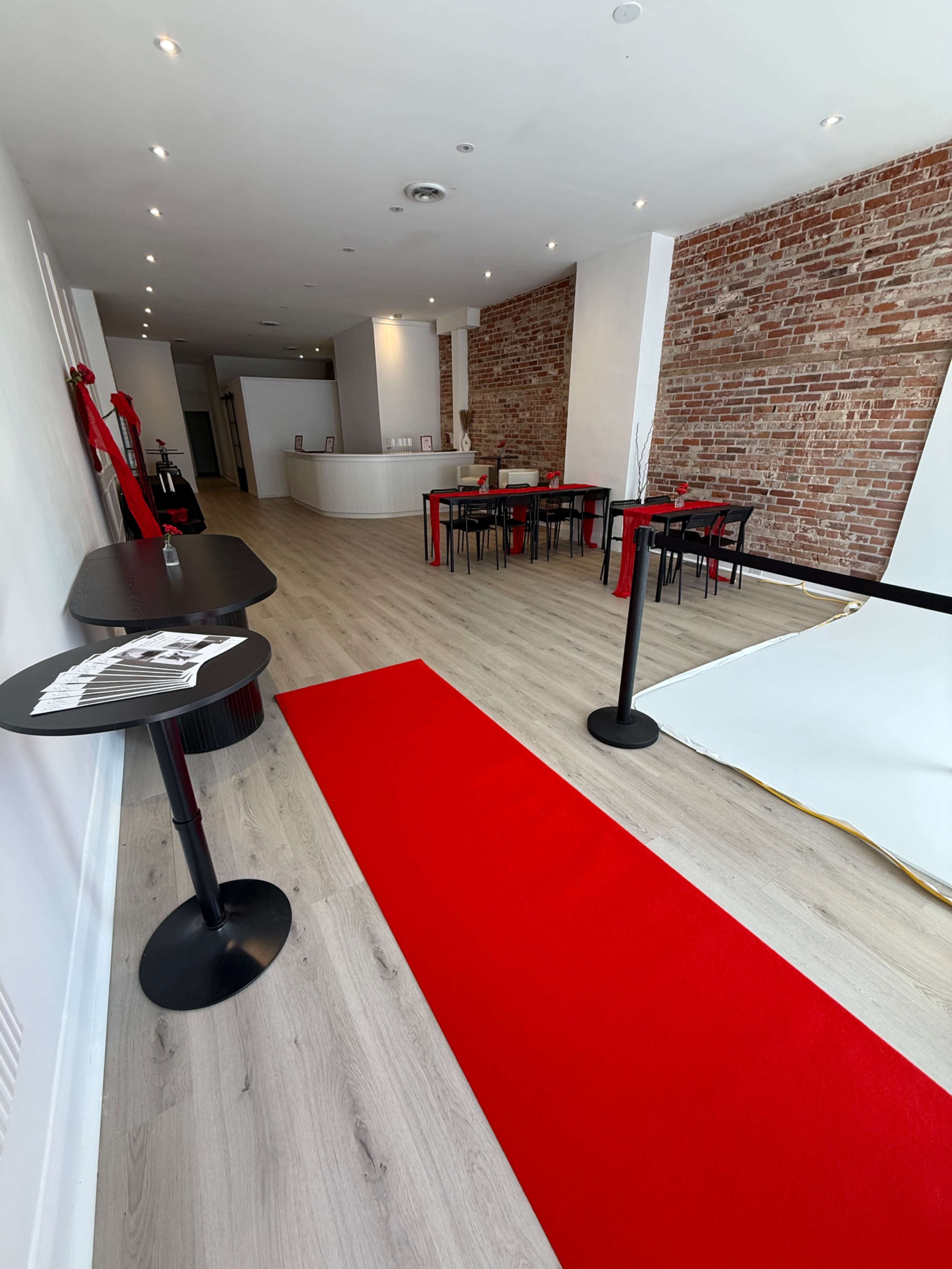 The image shows a modern, empty interior space with a red carpet, black tables, and a brick wall, illuminated by overhead lights.