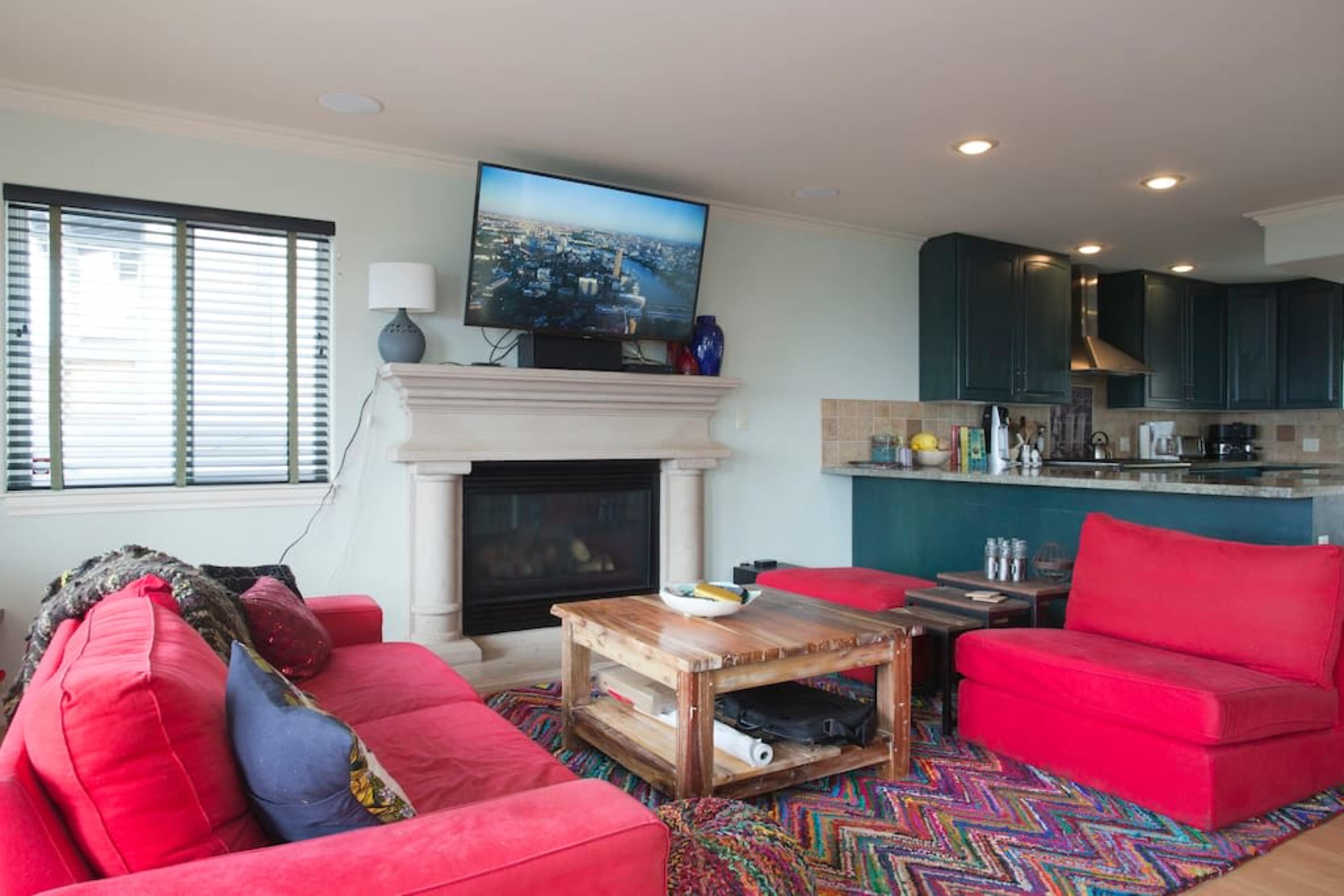 The image shows a living room with a red sofa set, a wooden coffee table, a fireplace, and a television mounted on the wall, with a kitchen visible in the background.