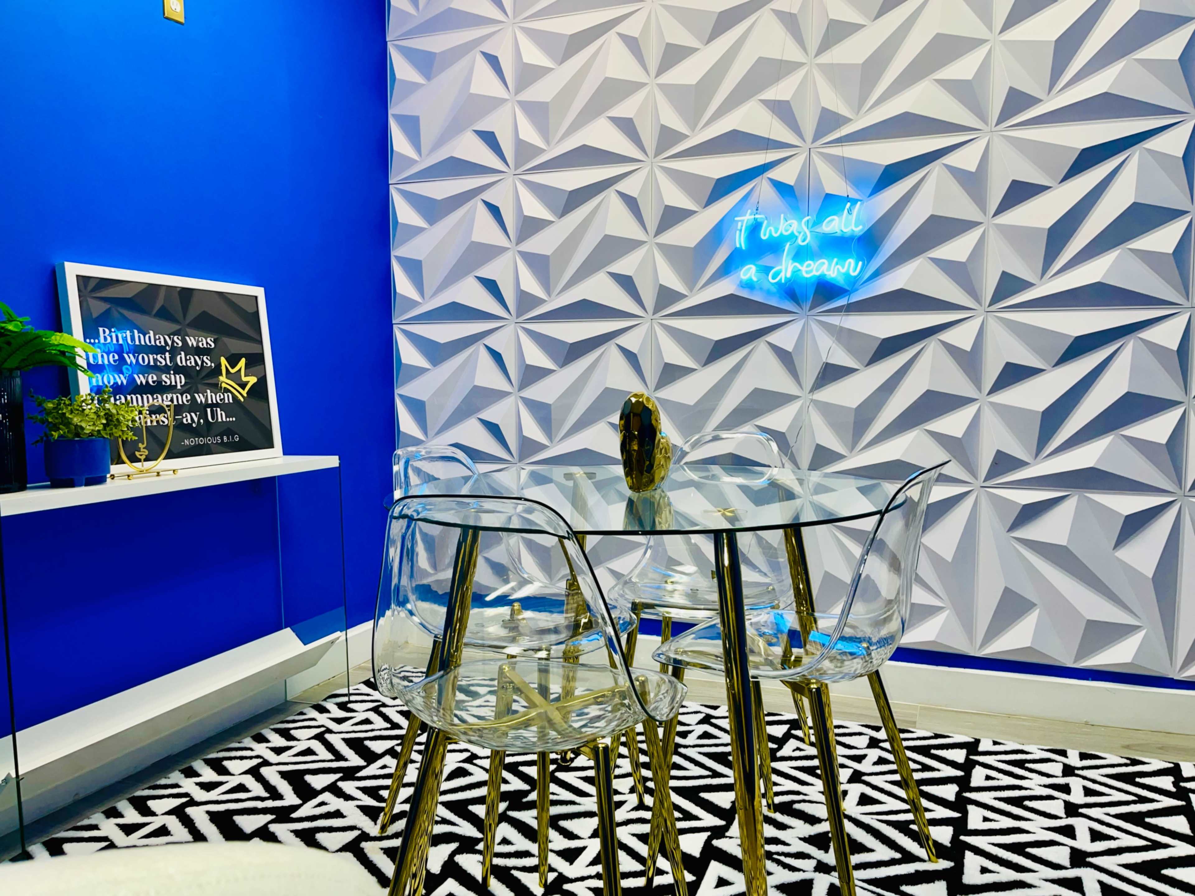 A modern dining area features a glass table with clear chairs, set against a blue wall adorned with geometric designs and a neon sign.