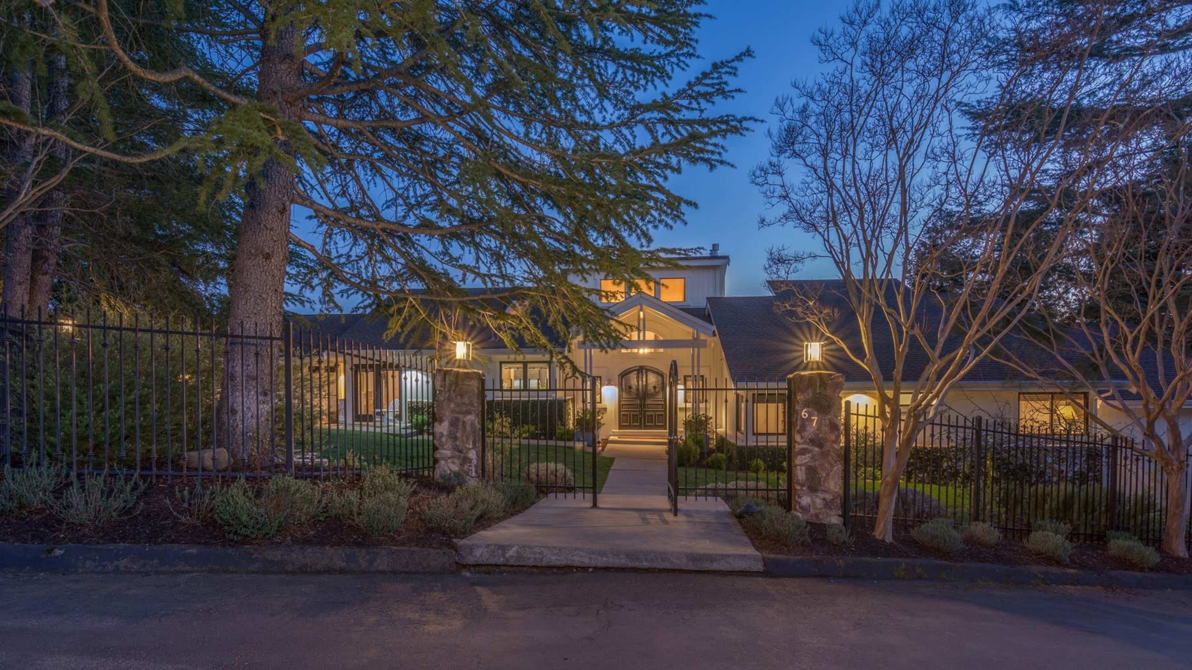 A well-lit, upscale house is framed by a gated entrance and surrounded by trees at dusk.