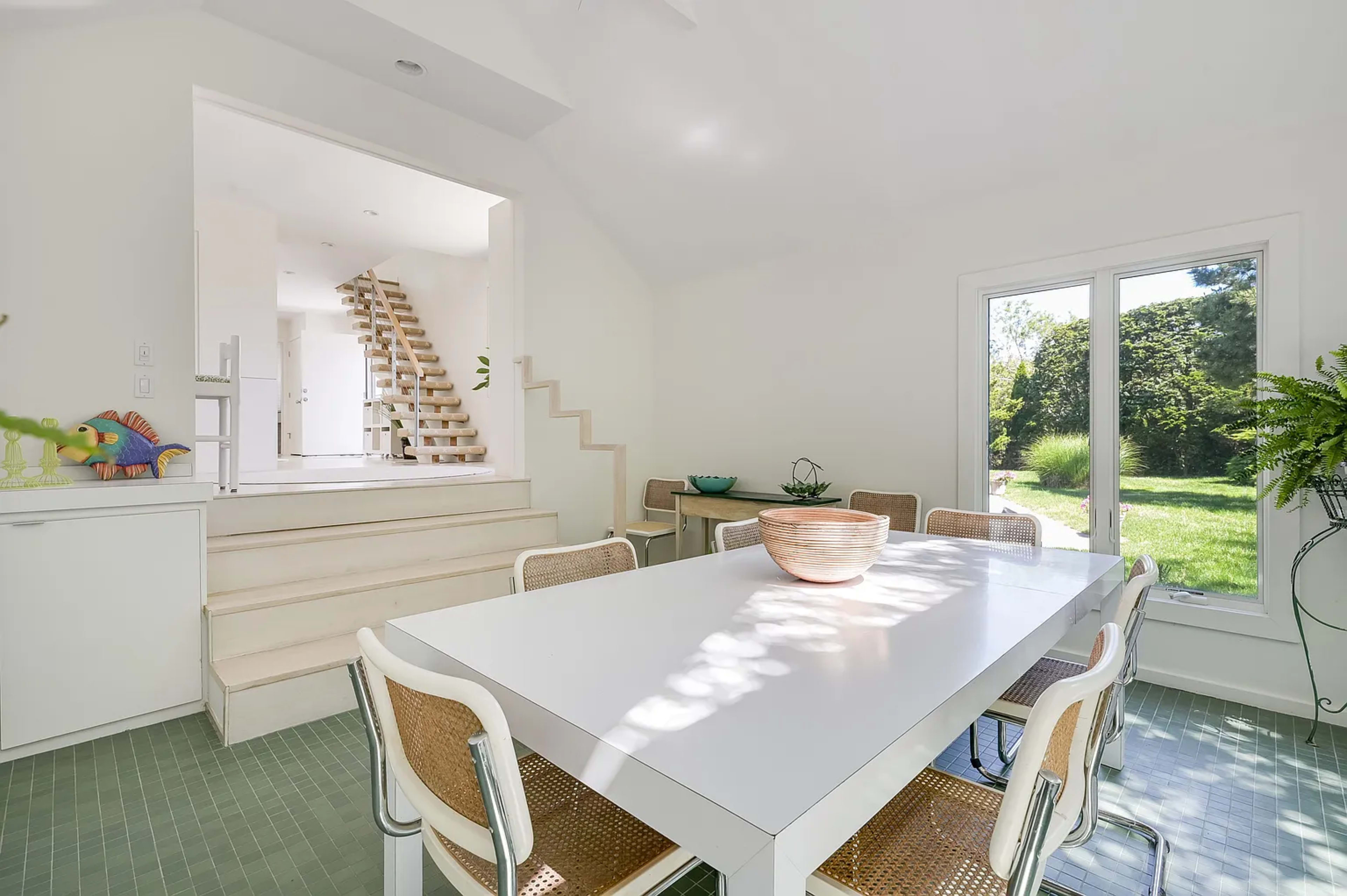 A modern dining area features a white table and chairs, with a staircase leading to a bright space beyond and large windows overlooking a garden.