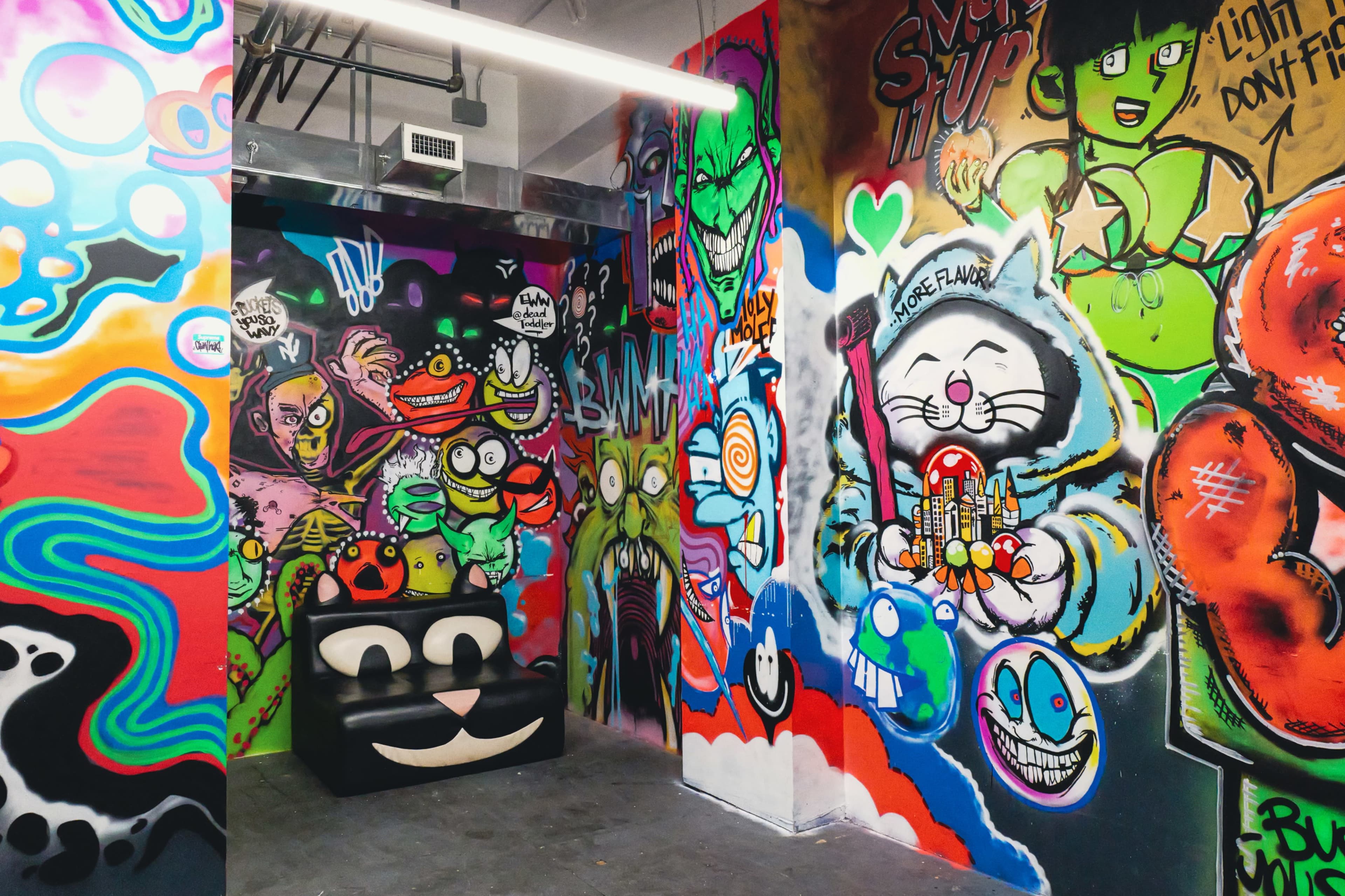 The image shows a vibrant room filled with colorful graffiti art on the walls, featuring various cartoon characters and designs, with a black couch in the center.