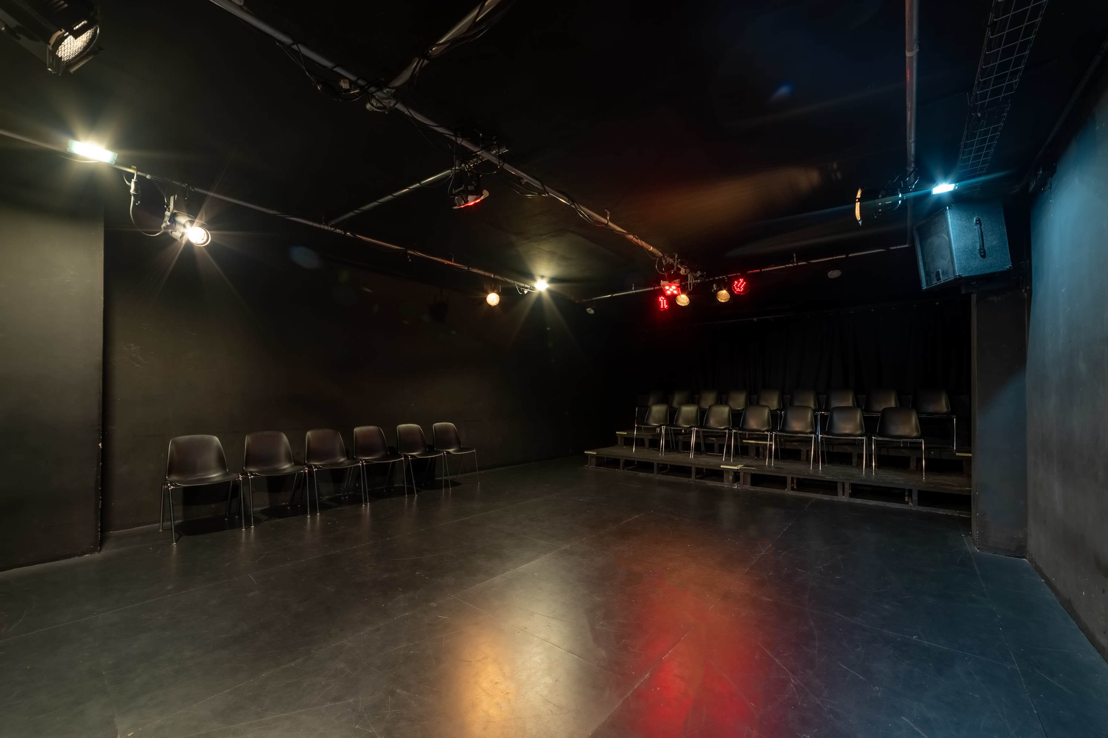 A dimly lit theater space with black walls, a stage, and rows of chairs arranged for an audience.
