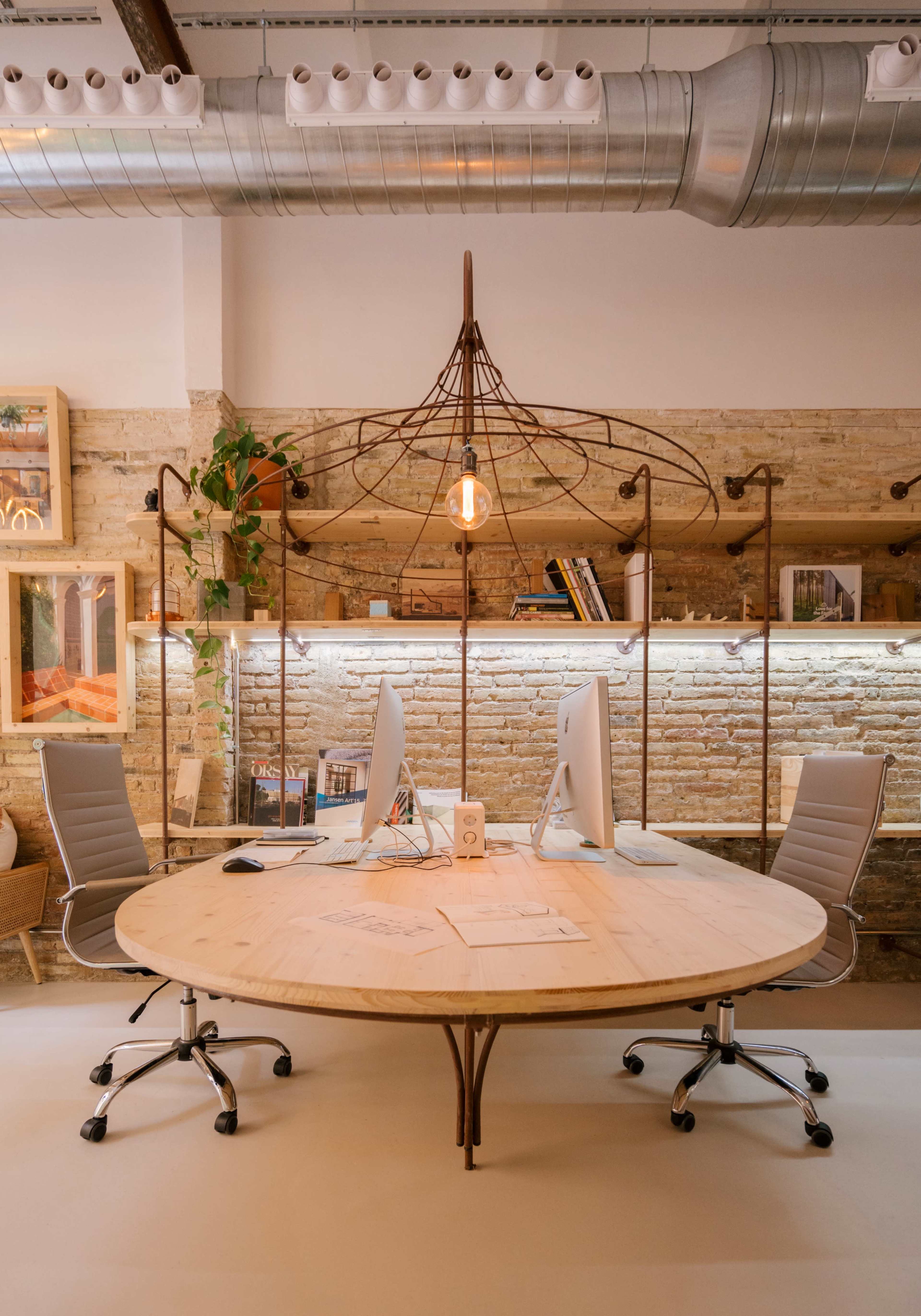 The image shows a modern office workspace featuring a round wooden table surrounded by two ergonomic chairs, with two computer monitors positioned at the table center and illuminated by a hanging light fixture.
