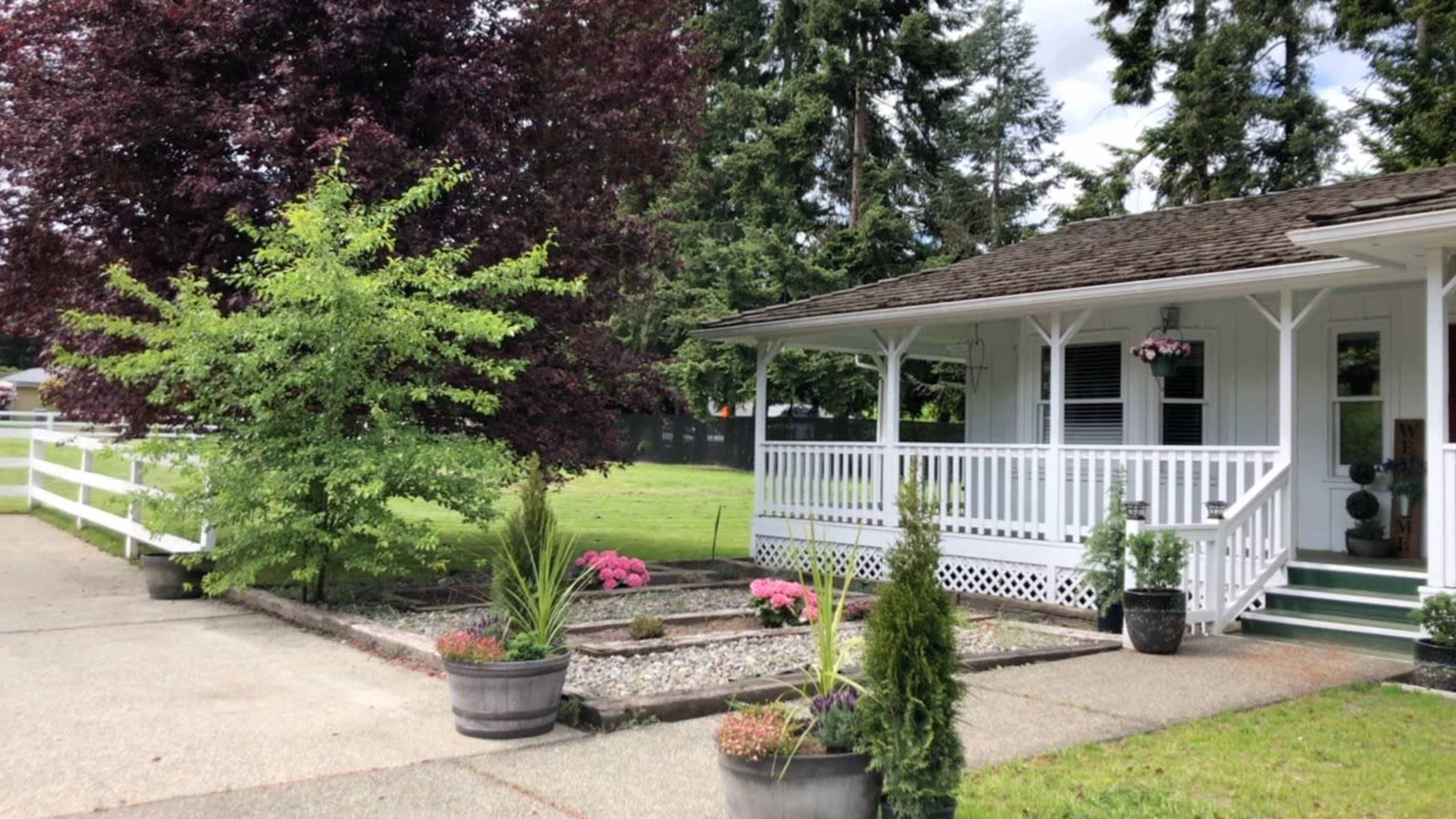 A welcoming house features a front porch with white railings, surrounded by landscaped garden beds and a driveway lined with decorative plants.