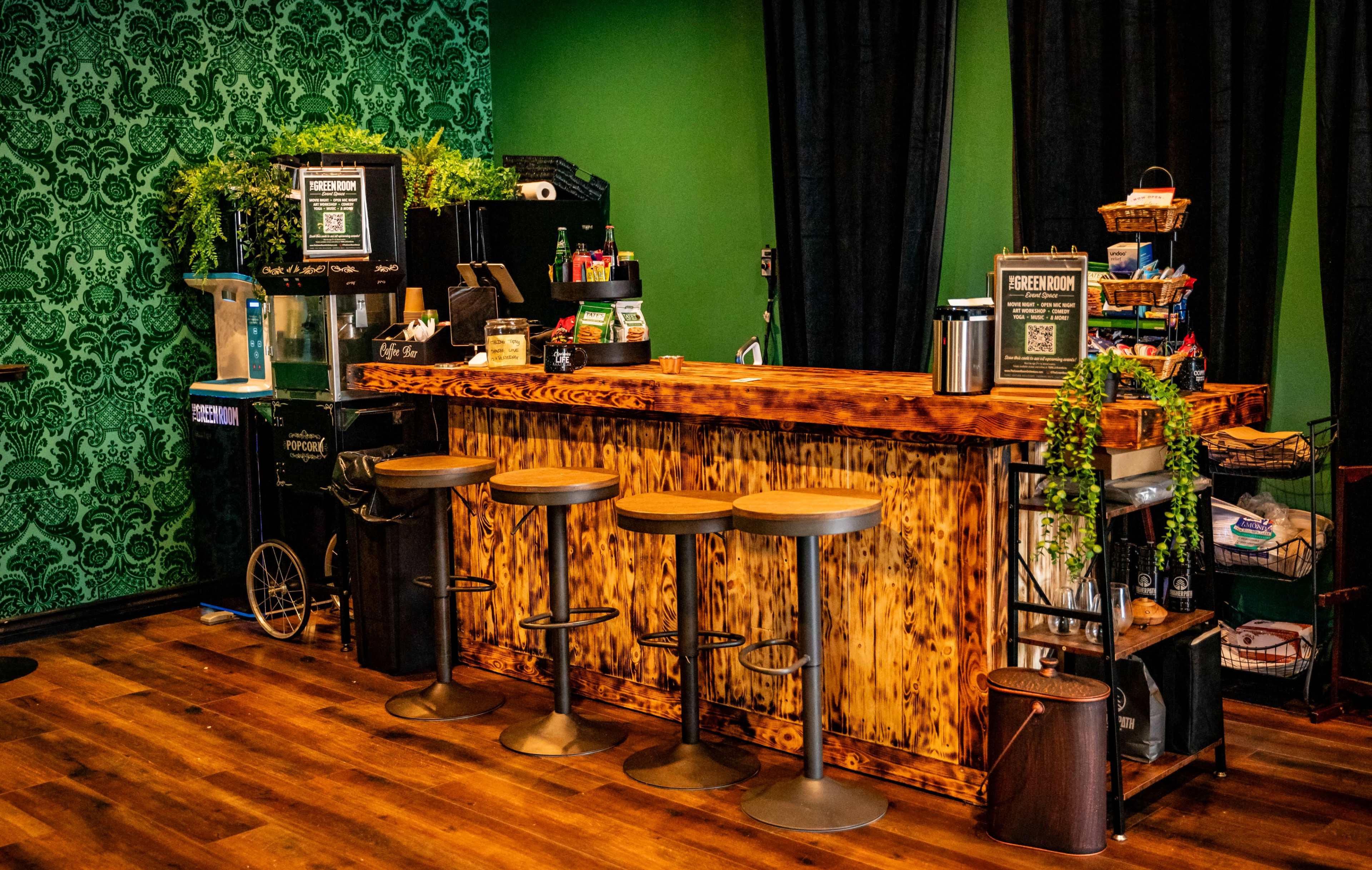 A coffee bar with a wooden counter and three metal stools, set against a green patterned wall.