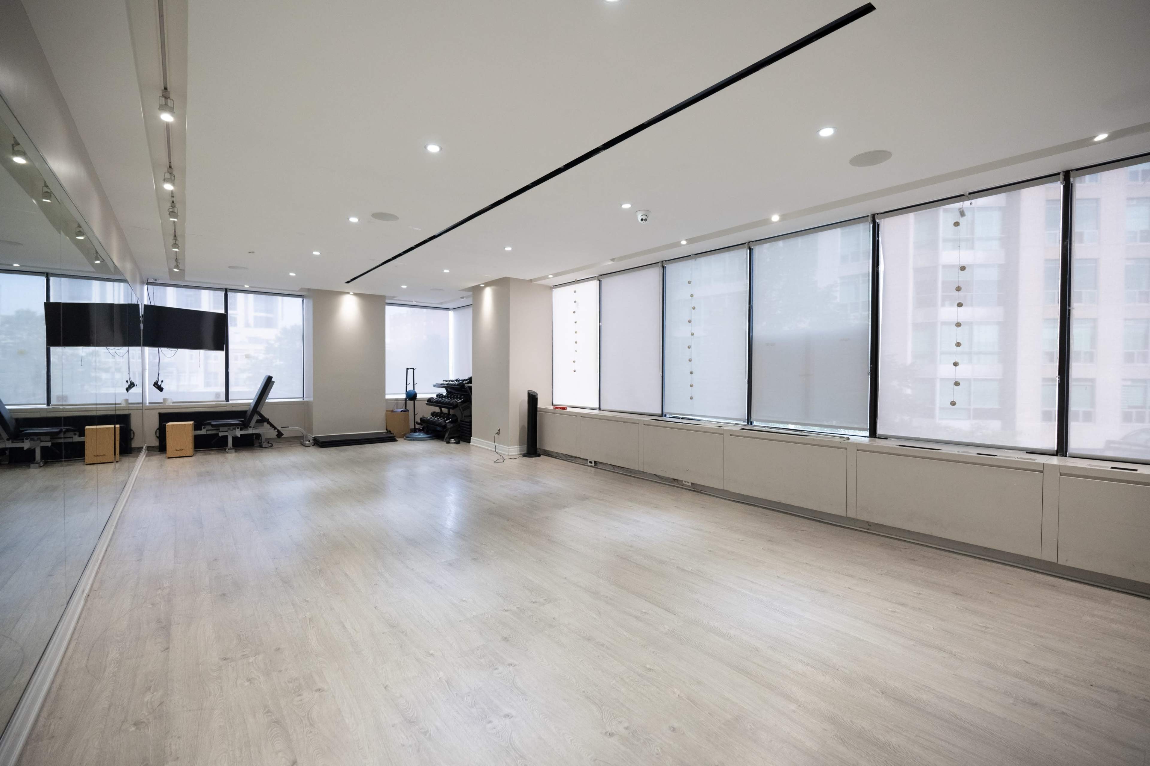 The image shows a spacious, empty room with light-colored floors, large windows, and a wall of mirrors on one side.