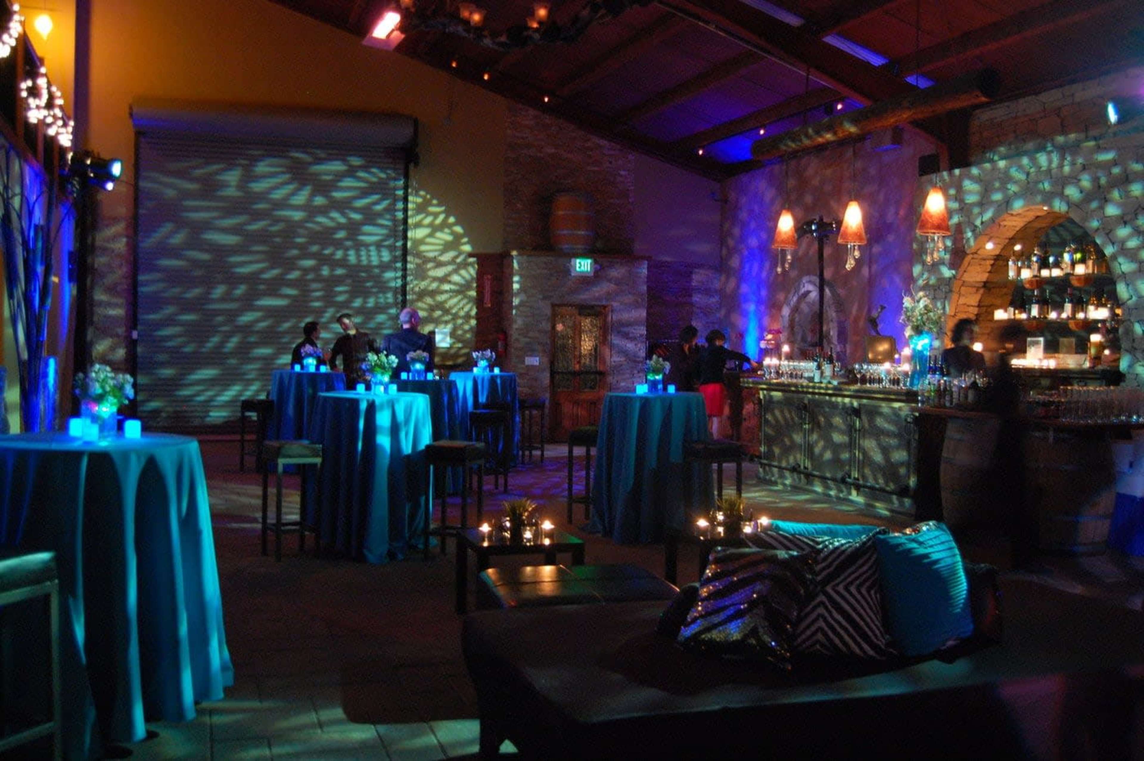 A dimly lit event space with high tables draped in blue cloth, a bar area in the background, and decorative lighting creating patterns on the walls.