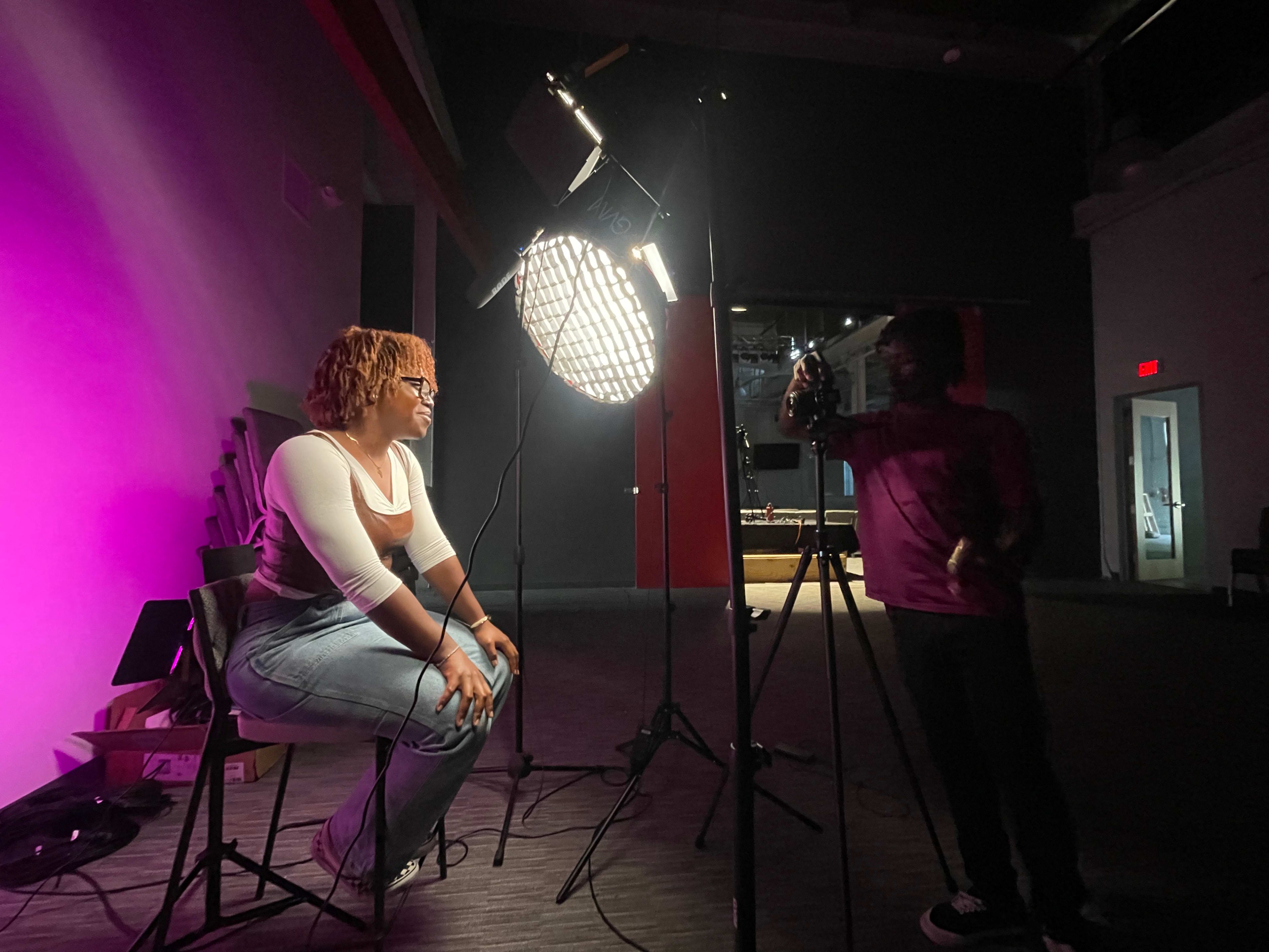 A person with curly hair sits on a stool, illuminated by a large light source, while another individual stands in the shadows holding a camera.