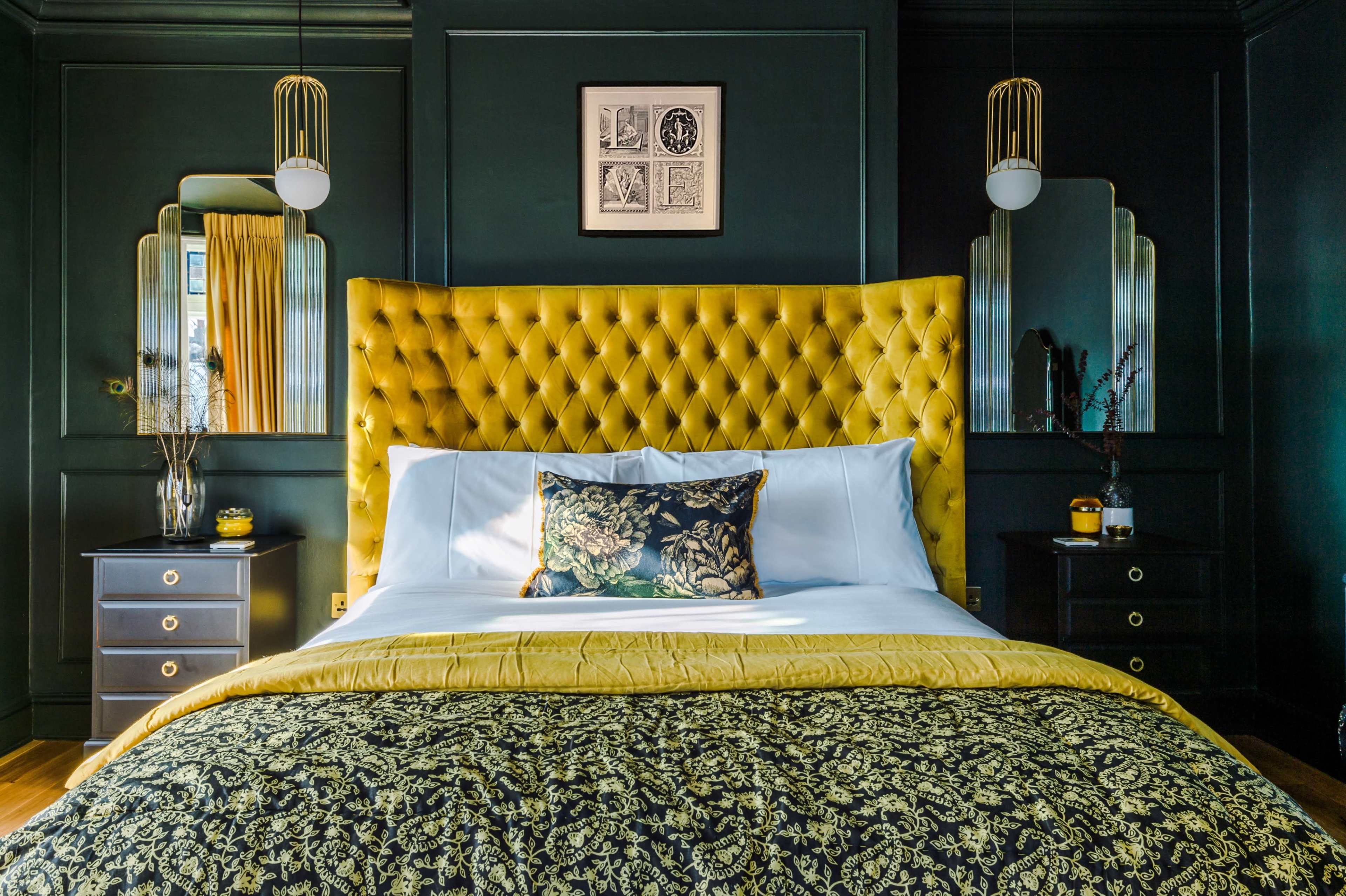 A bedroom features a large yellow tufted headboard, a black and gold patterned bedspread, and dark green walls with stylish lighting fixtures.