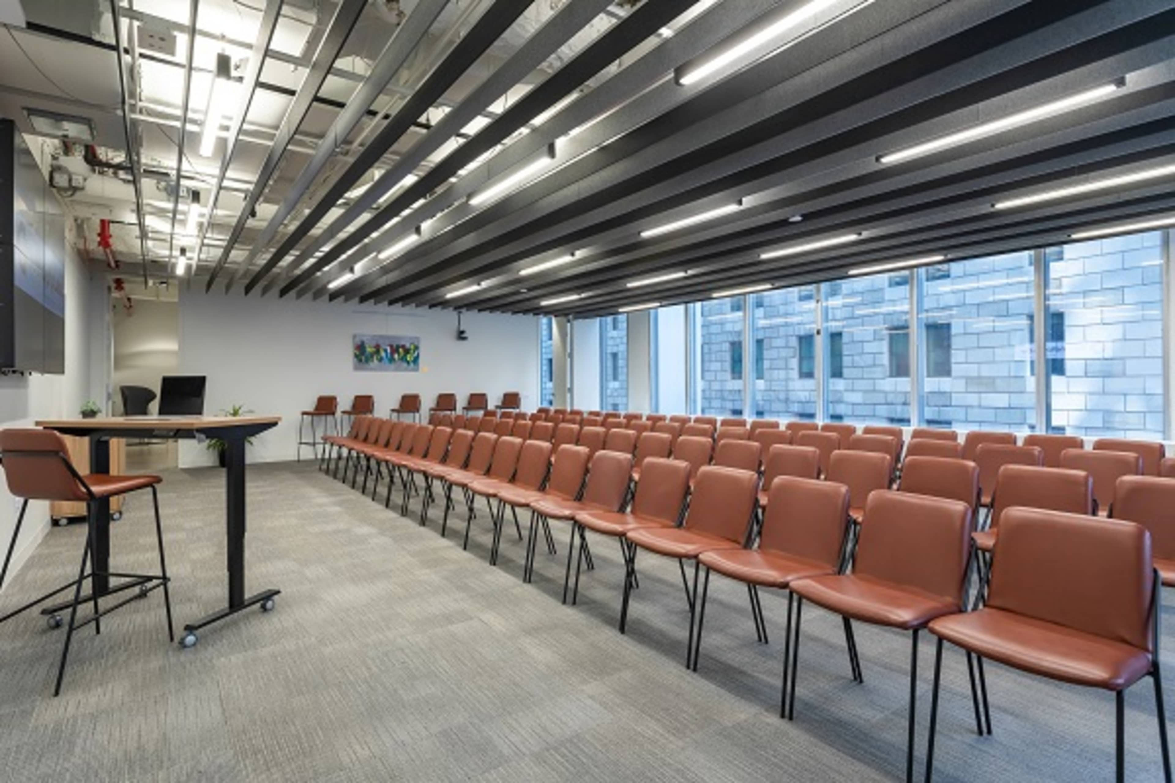 A modern conference room features rows of brown chairs facing a presentation area, with large windows casting natural light across the space.