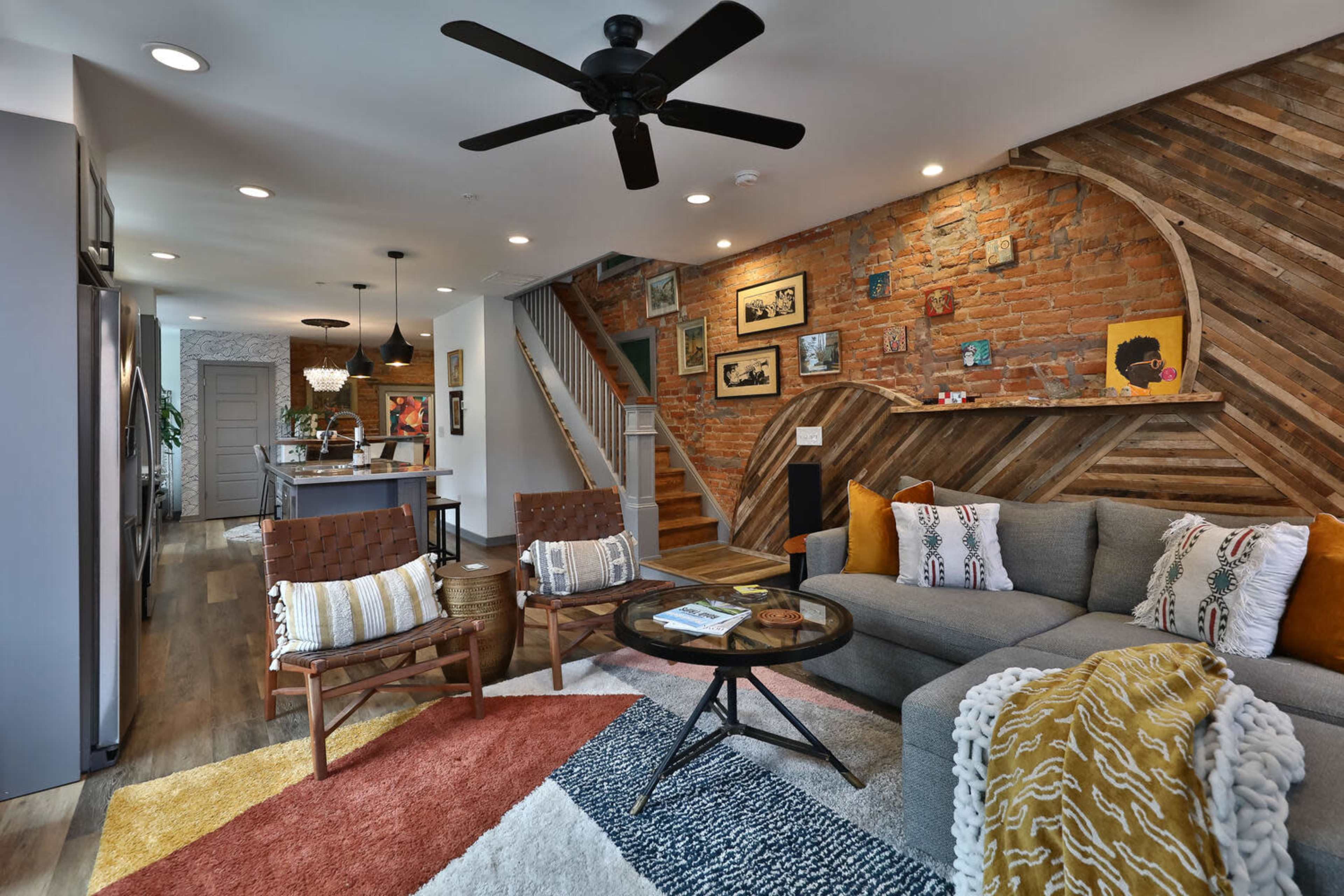 A modern living room featuring a gray sofa, wooden accents, a coffee table, and a staircase in the background, with a brick wall and colorful rugs.