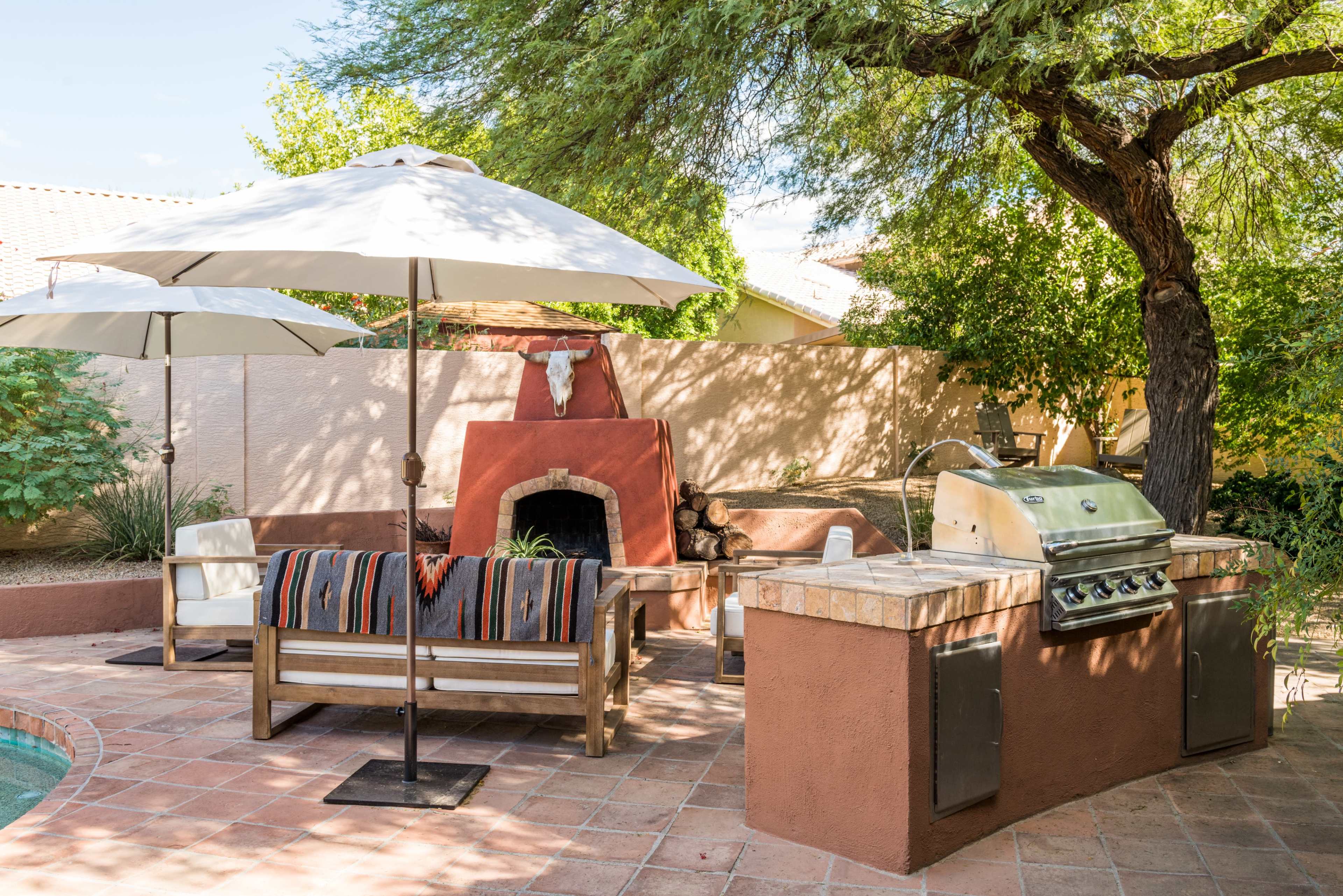 The image shows a backyard patio featuring a grill, a stone fireplace with a decorative element, outdoor seating under umbrellas, and a pool.