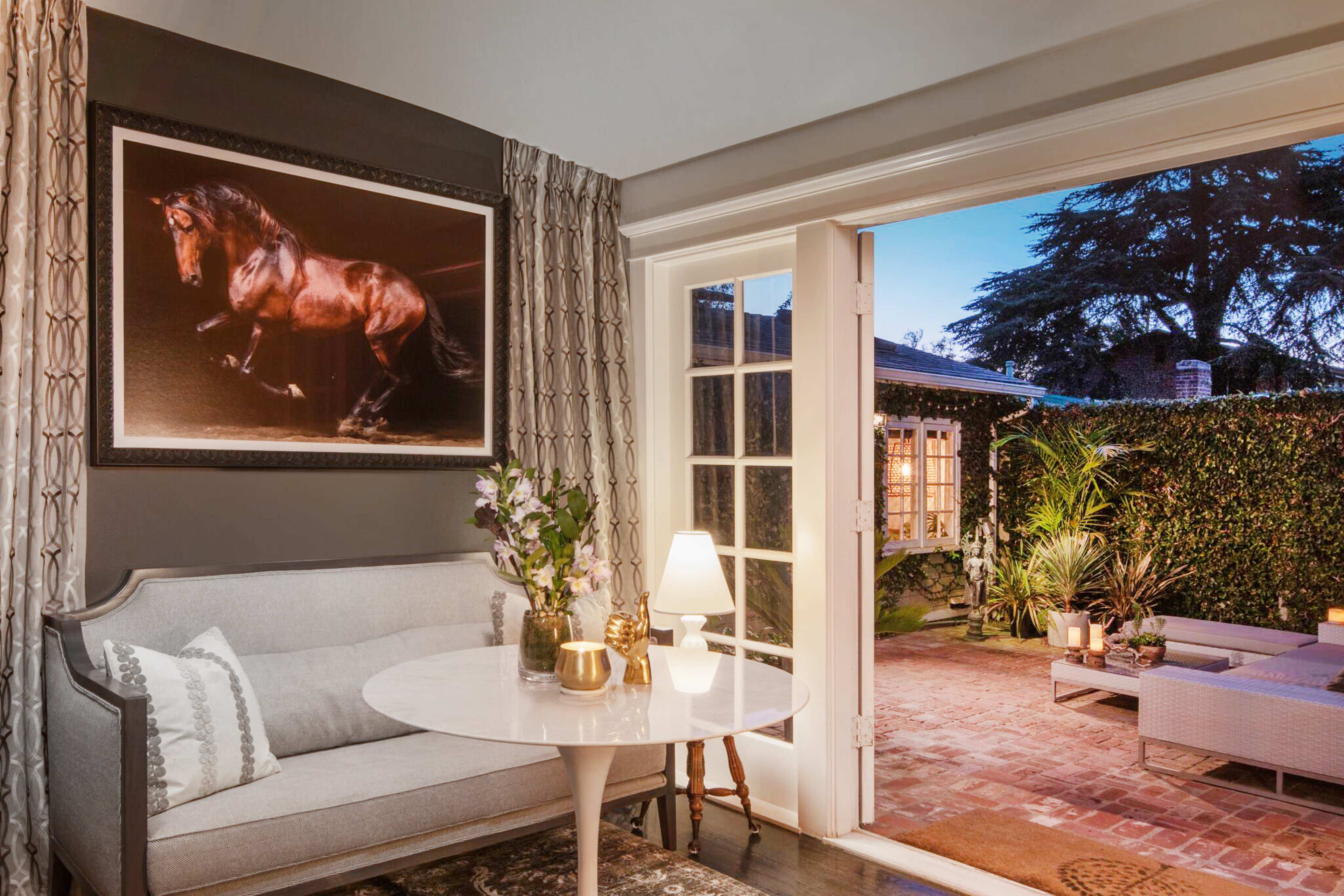 A cozy corner features a gray sofa and a small round table, with a large horse painting on the wall and French doors opening to a brick patio.