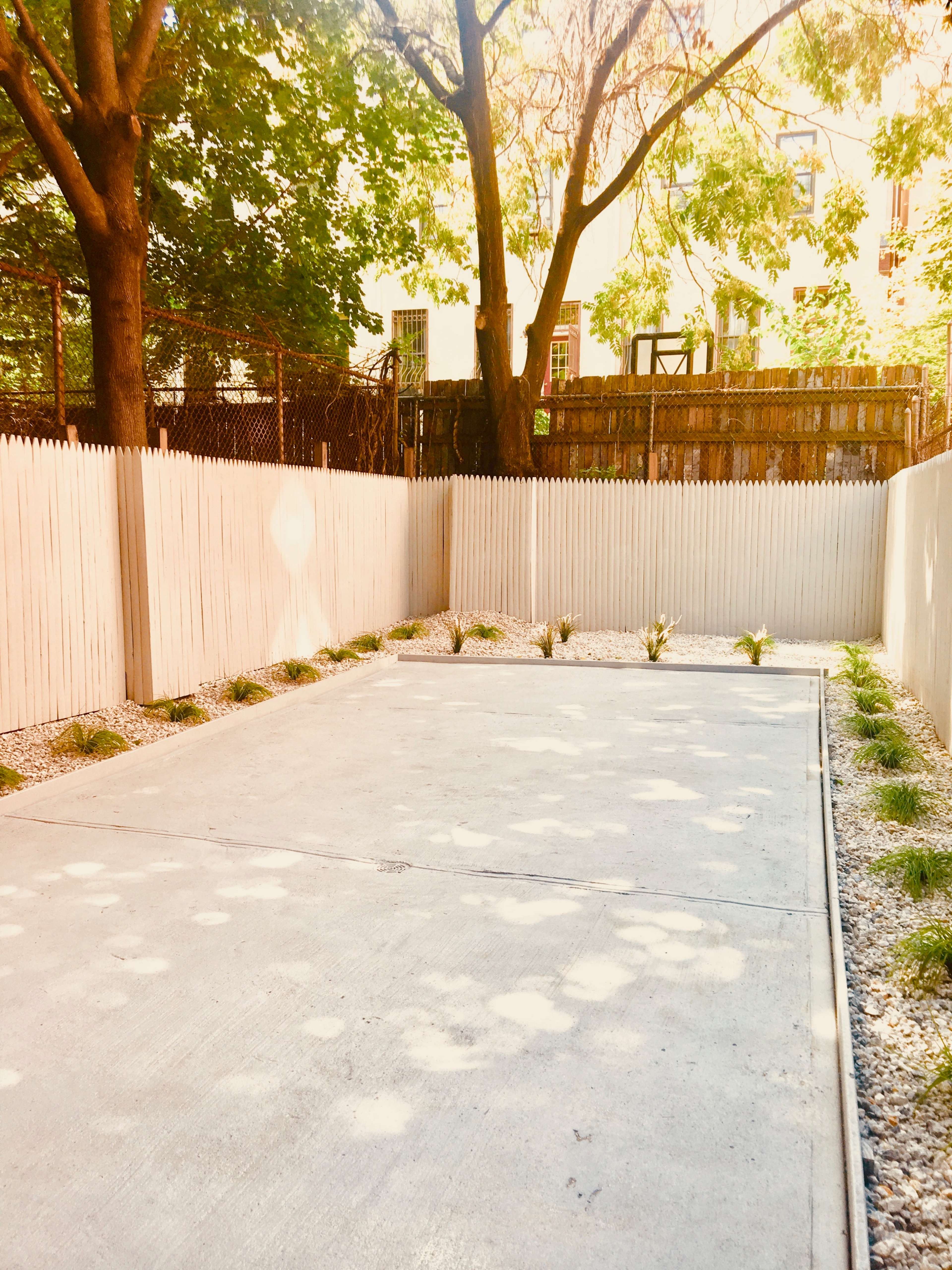A concrete outdoor space is bordered by a white picket fence and features small plants along the edges.