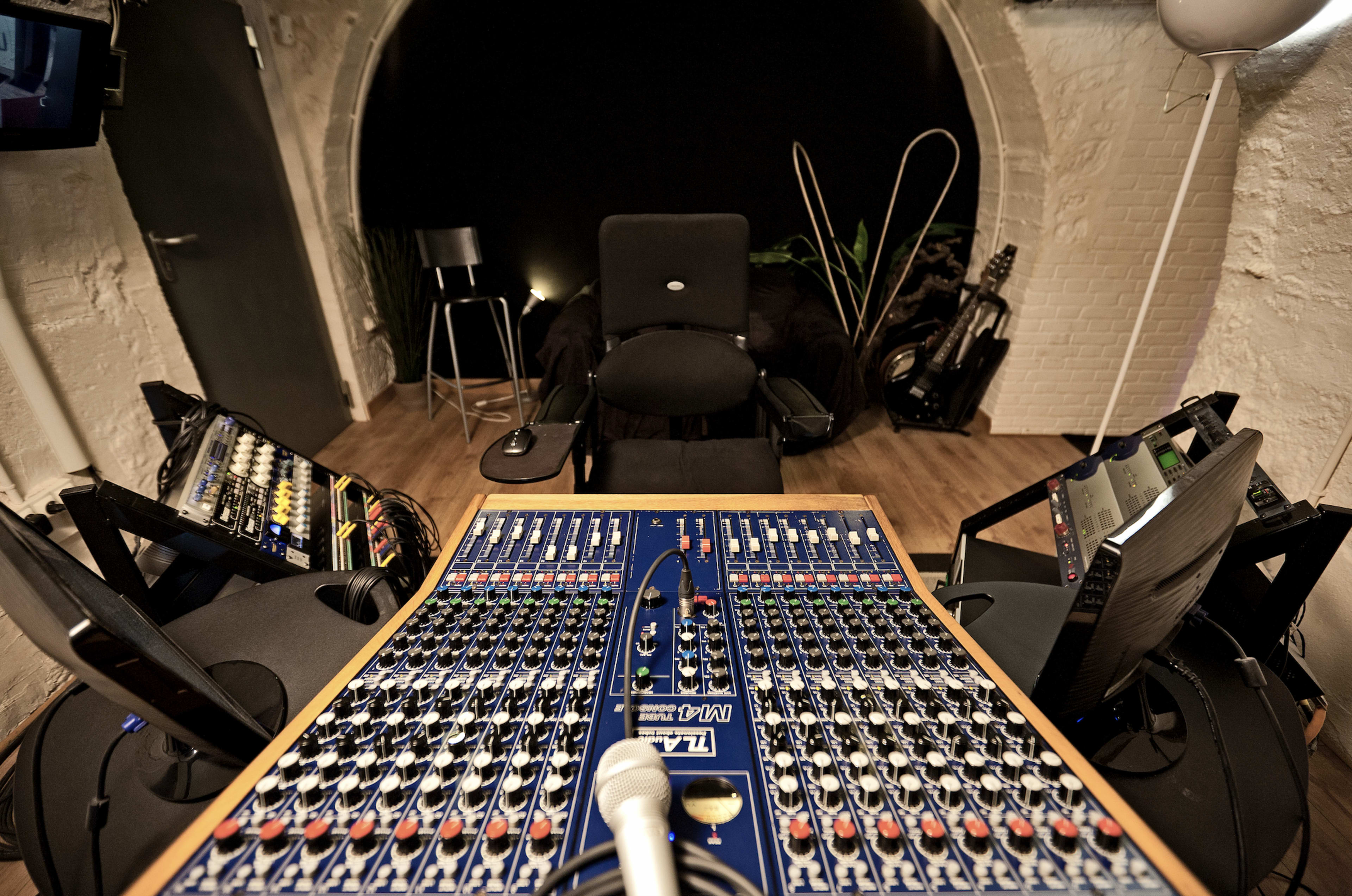The image shows a sound mixing studio with a large mixing console at the center, flanked by two monitors, and a microphone positioned in front of a black chair.