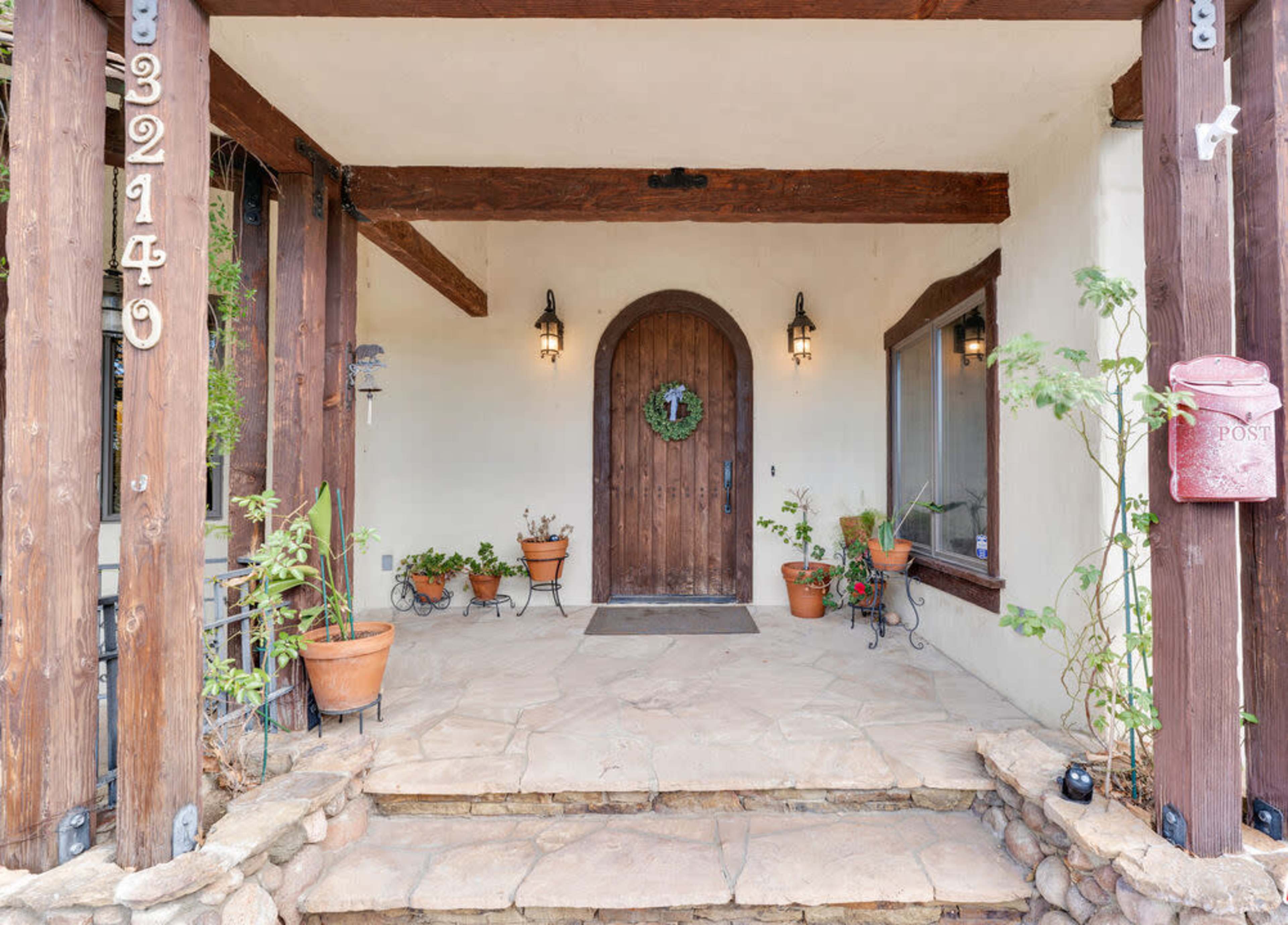The image shows a entrance with a wooden door surrounded by potted plants and stone steps.