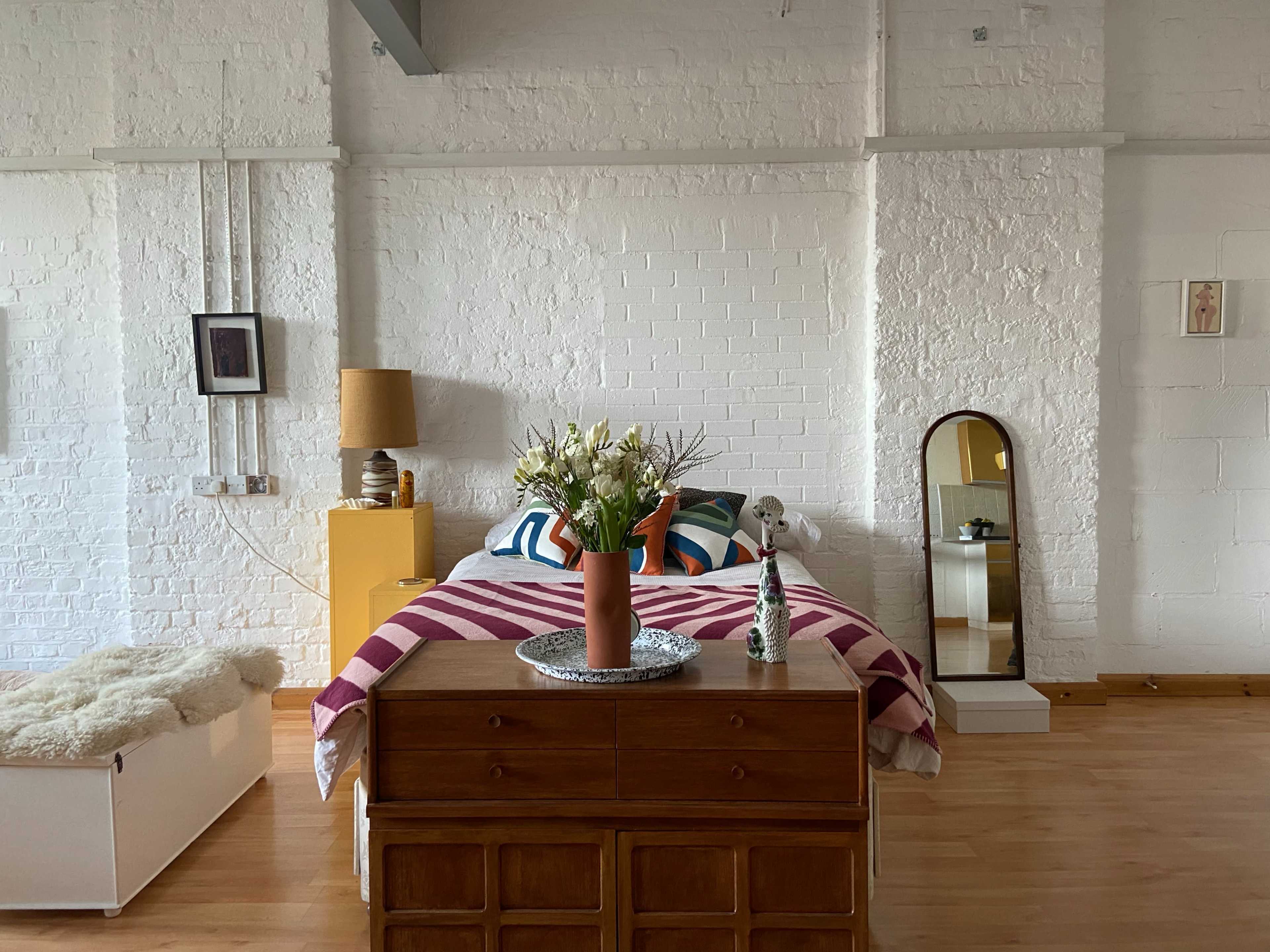 A minimalist bedroom with a wooden dresser in front of a bed adorned with a striped blanket, flanked by a vase of flowers and a mirror.
