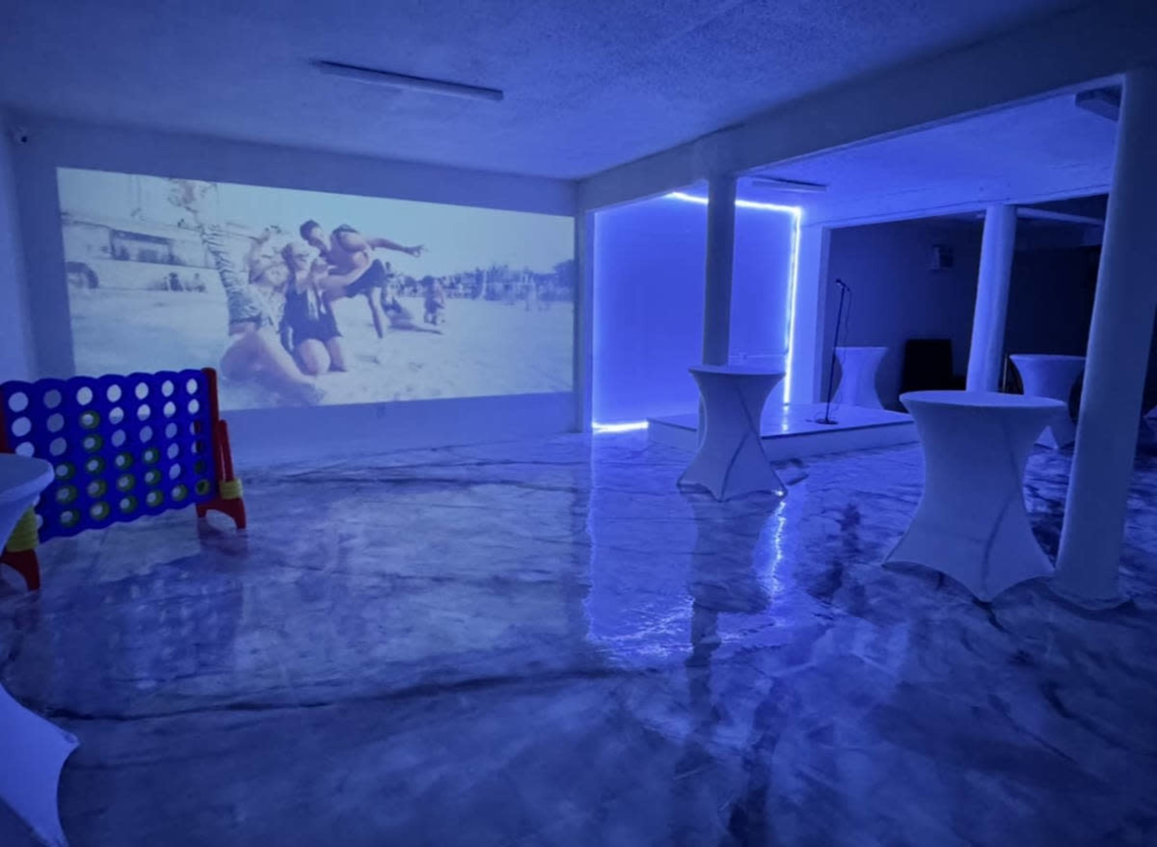 The image shows an indoor space with a projector displaying a video on one wall, illuminated by blue LED lighting and featuring white tables arranged around a reflective floor.