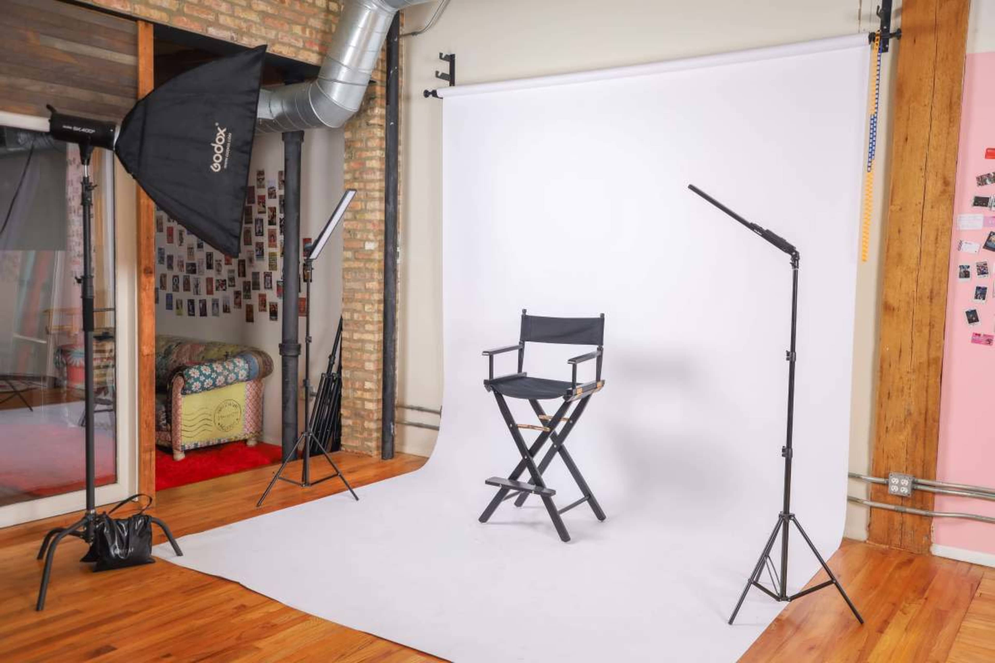 The image shows a photography studio setup with a black director's chair, three light sources, and a white backdrop.
