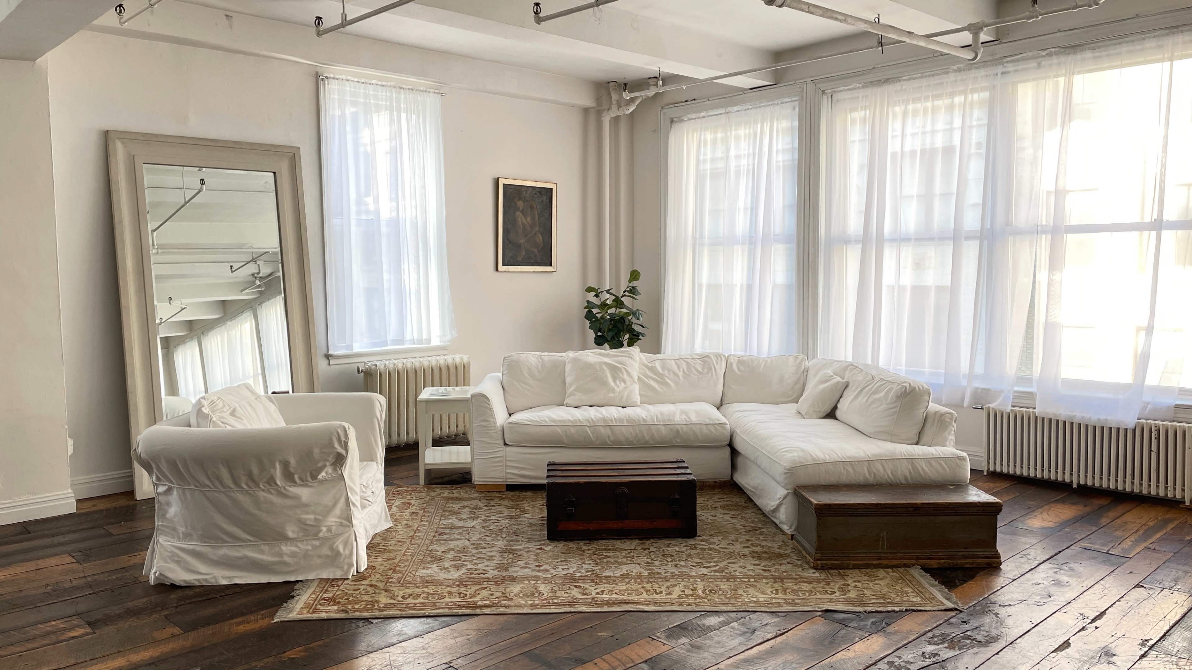 A sunlit room with a white sectional sofa, a matching armchair, a wooden coffee table, and large windows draped with sheer curtains.