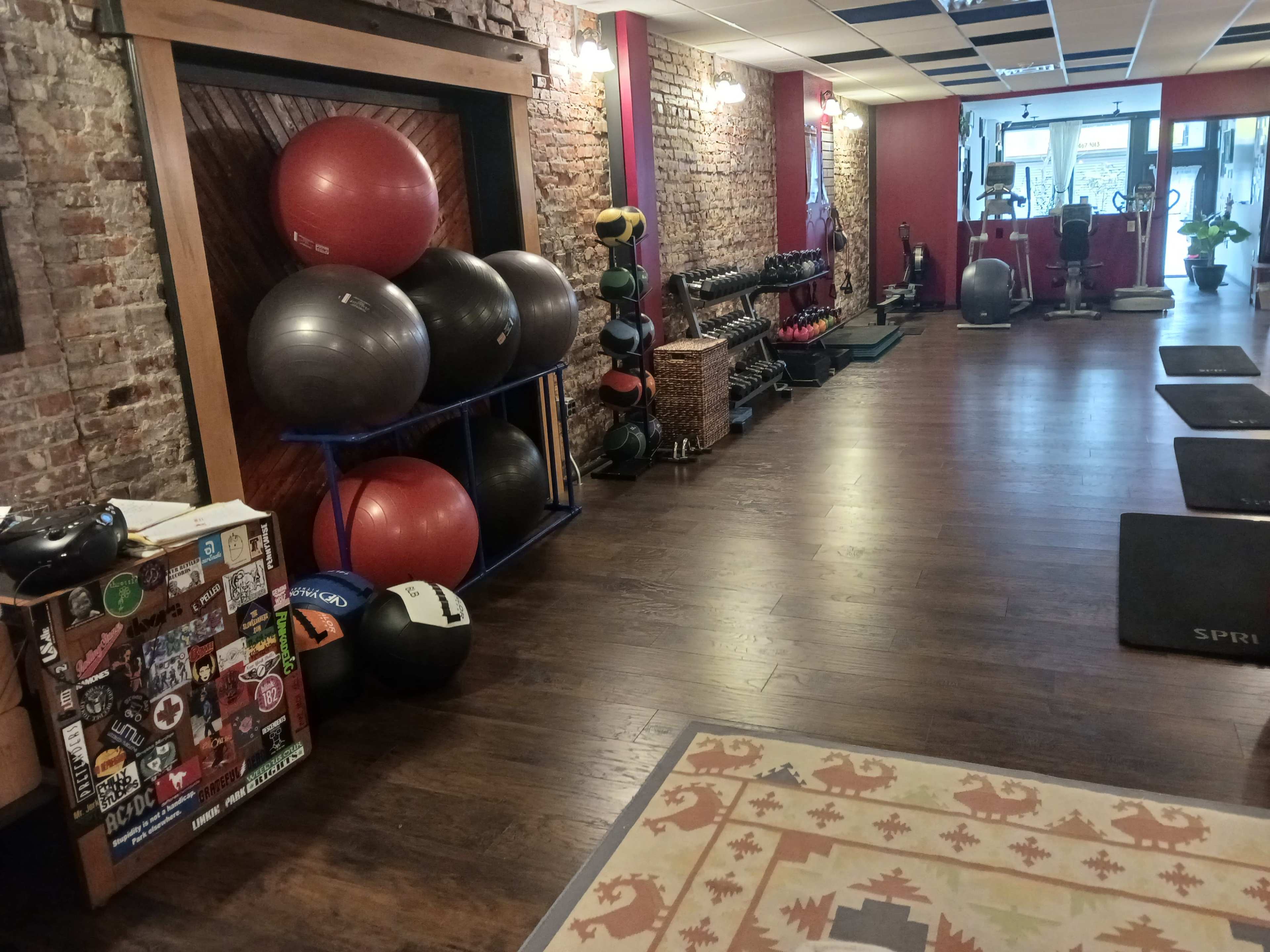 The image shows a gym interior with wooden flooring, brick walls, and various fitness equipment, including weights and exercise balls, along one side.