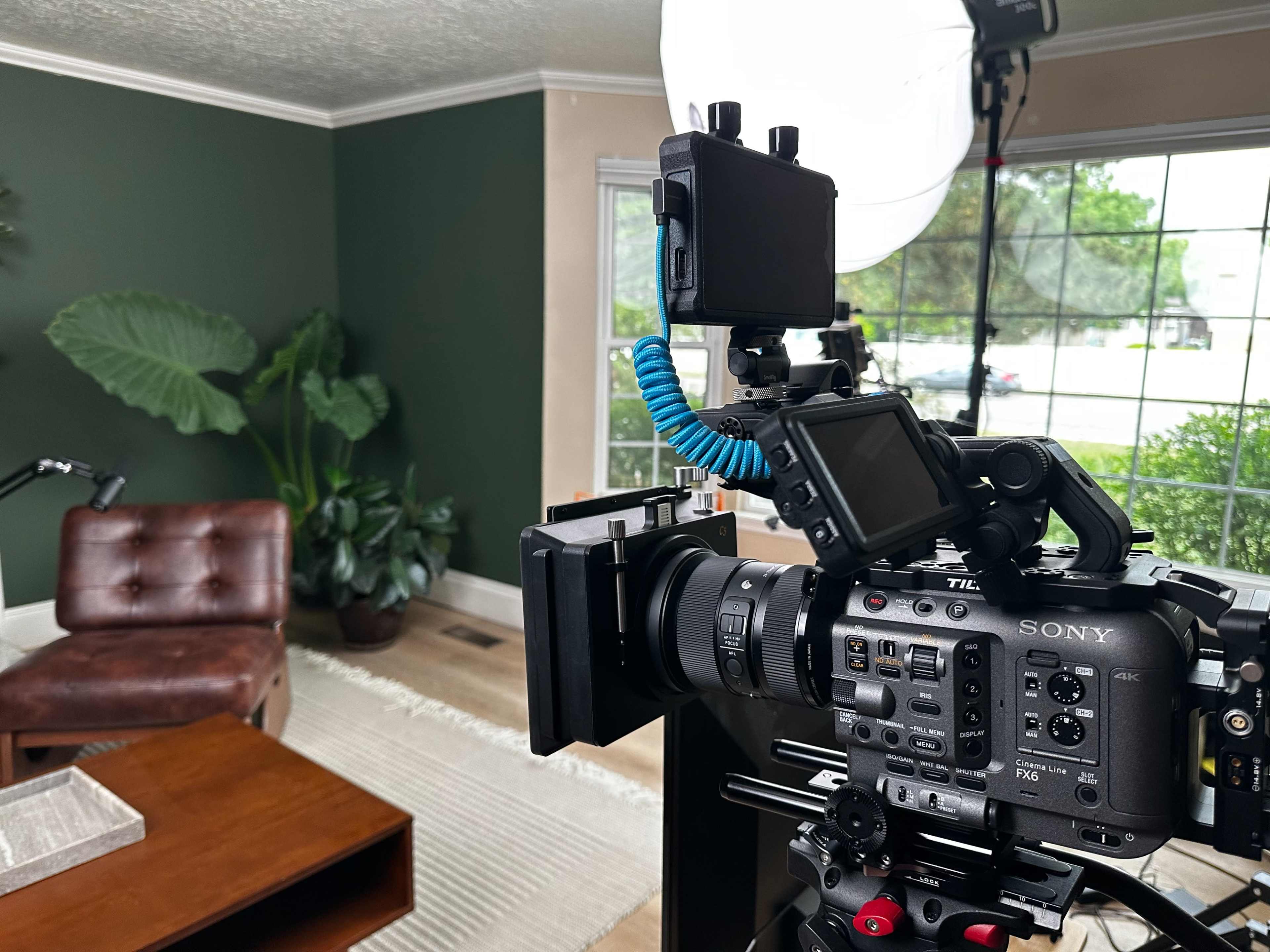 A camera is positioned on a tripod in a well-lit living room featuring a green accent wall, a brown leather chair, and several indoor plants.
