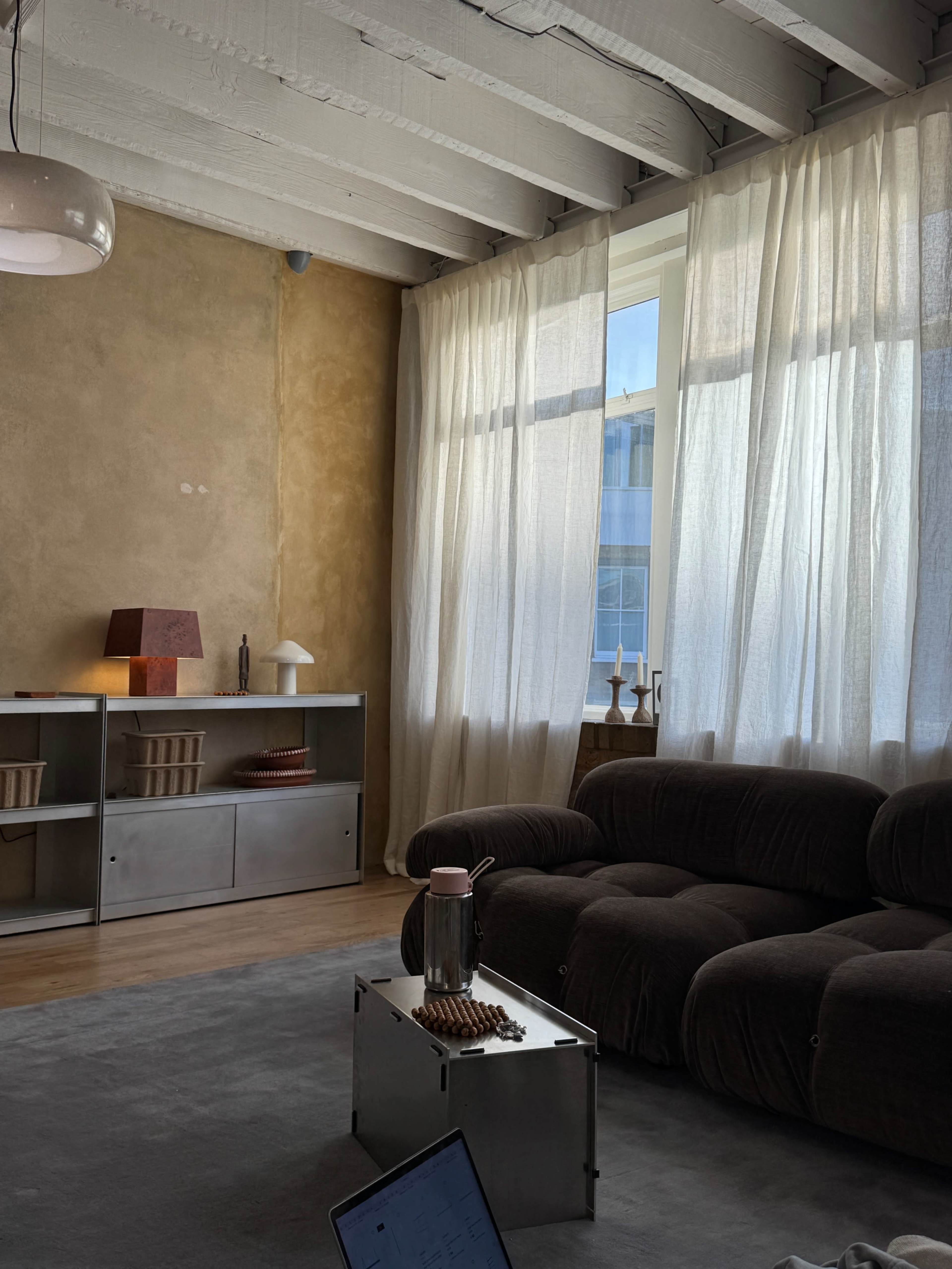 The image features a spacious living room with a brown sofa, a light-colored rug, sheer curtains letting in natural light, and a minimalist shelving unit with decorative items.
