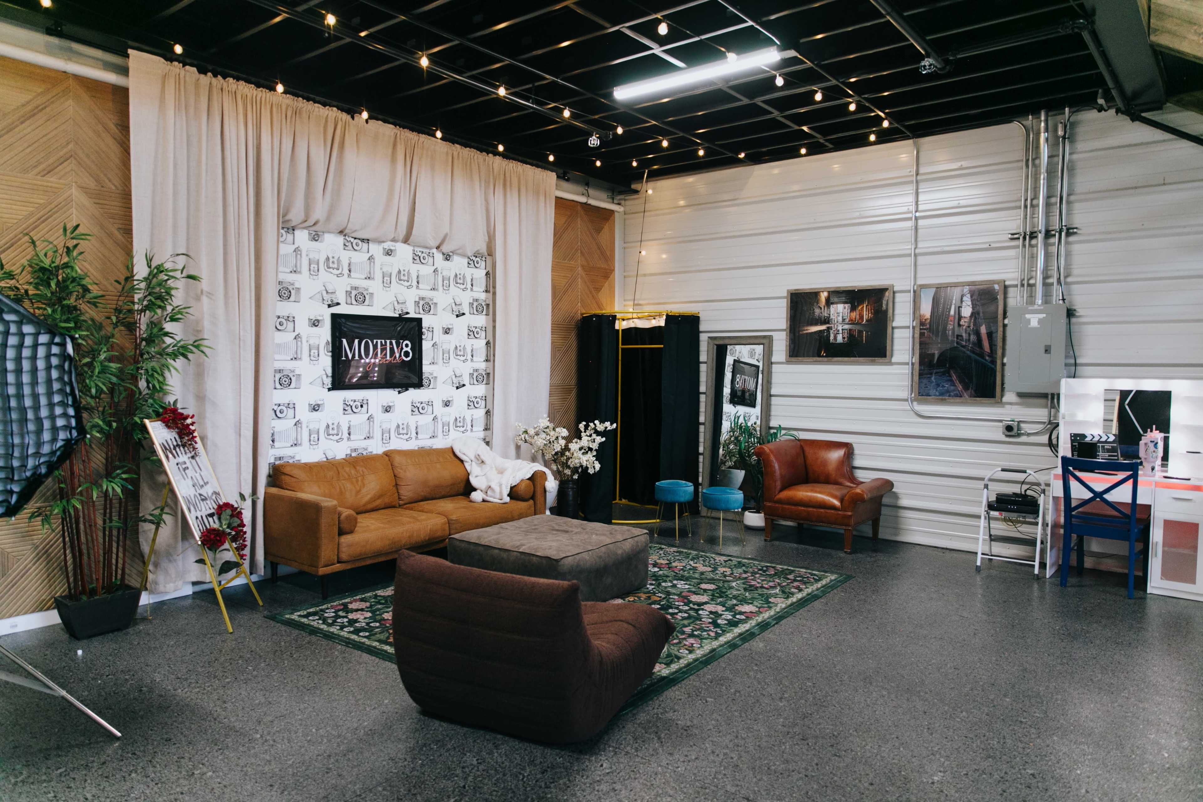 The image shows a modern studio space featuring a brown leather sofa, a gray seating arrangement, decorative plants, and a backdrop with a patterned wall.