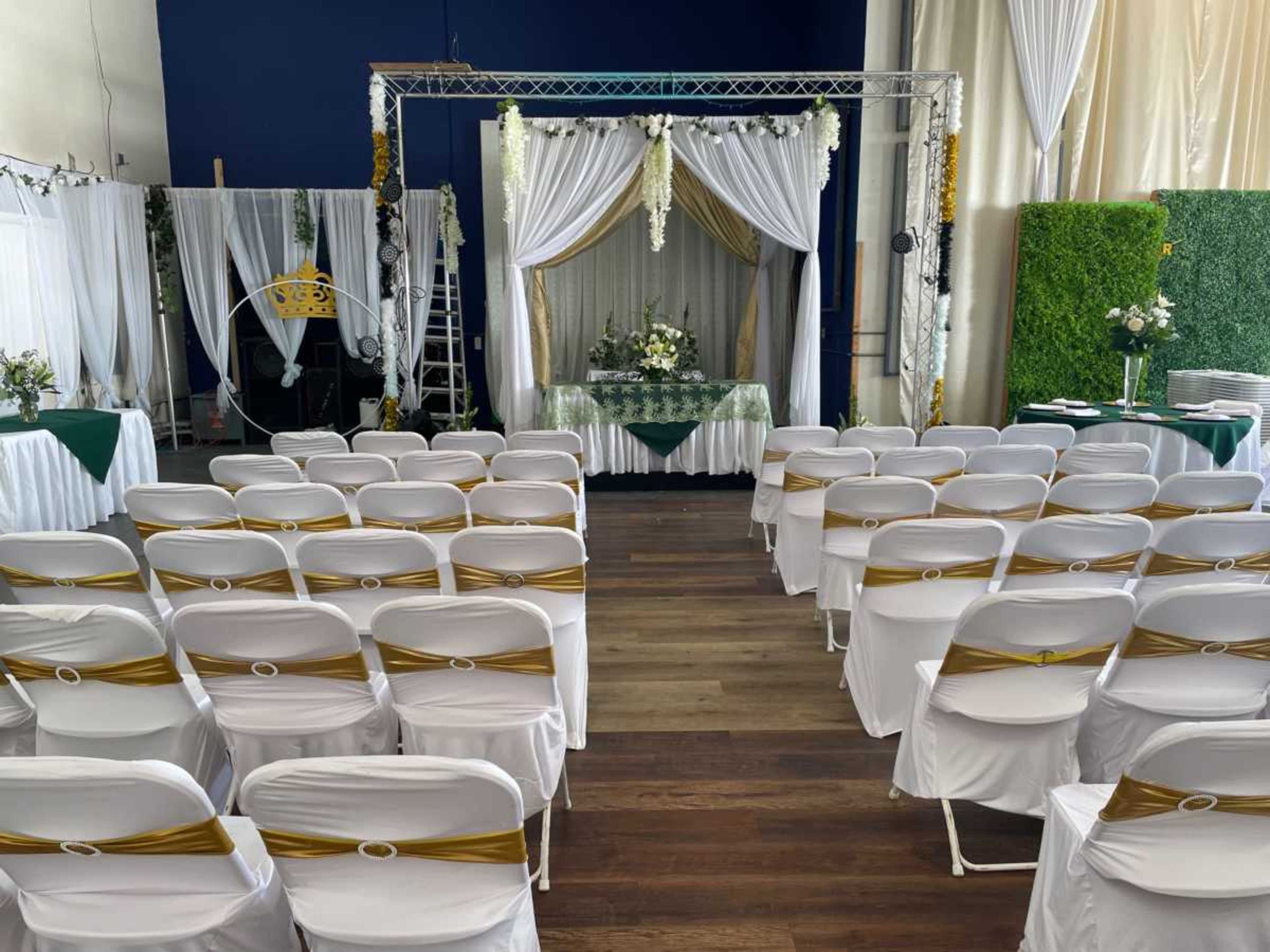 The image shows a decorated indoor venue with rows of white chairs facing an elegant altar draped in white fabric and adorned with flowers.