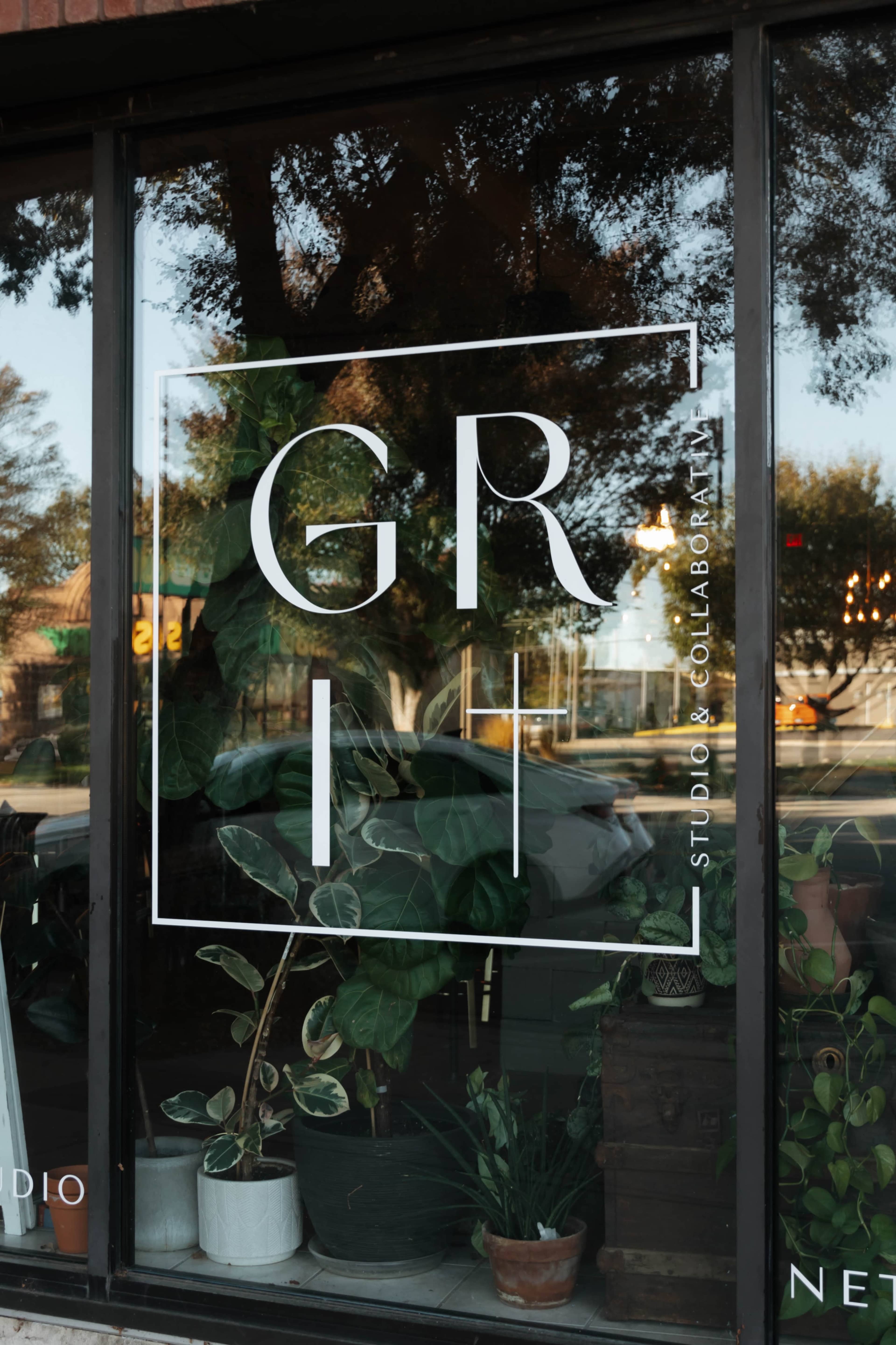 The image shows a storefront window displaying the word "GRIT" in large letters, along with the text "STUDIO COLLABORATIVE," and various potted plants visible inside.