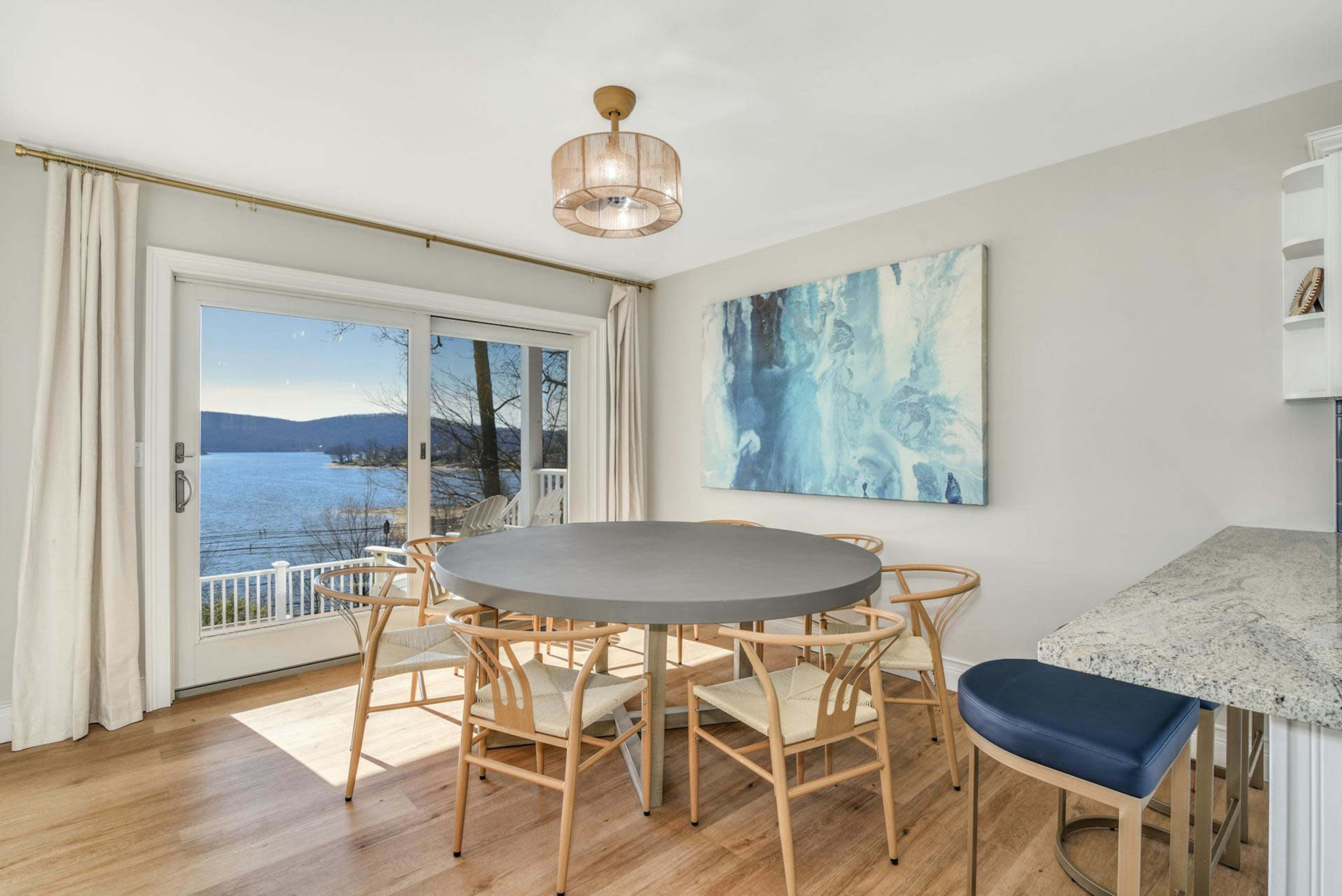 A circular dining table surrounded by chairs is positioned in a room with large windows overlooking a lake and mountains.